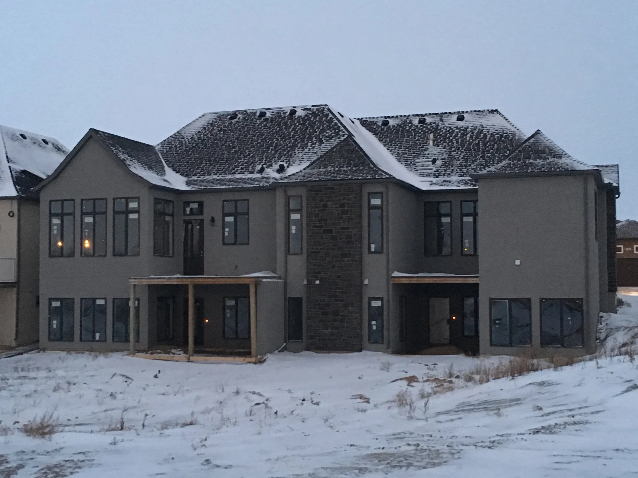 A residential house with a sloped roof covered in snow, large windows, and a wood deck in the backyard. The ground is snowy with patches of grass visible.