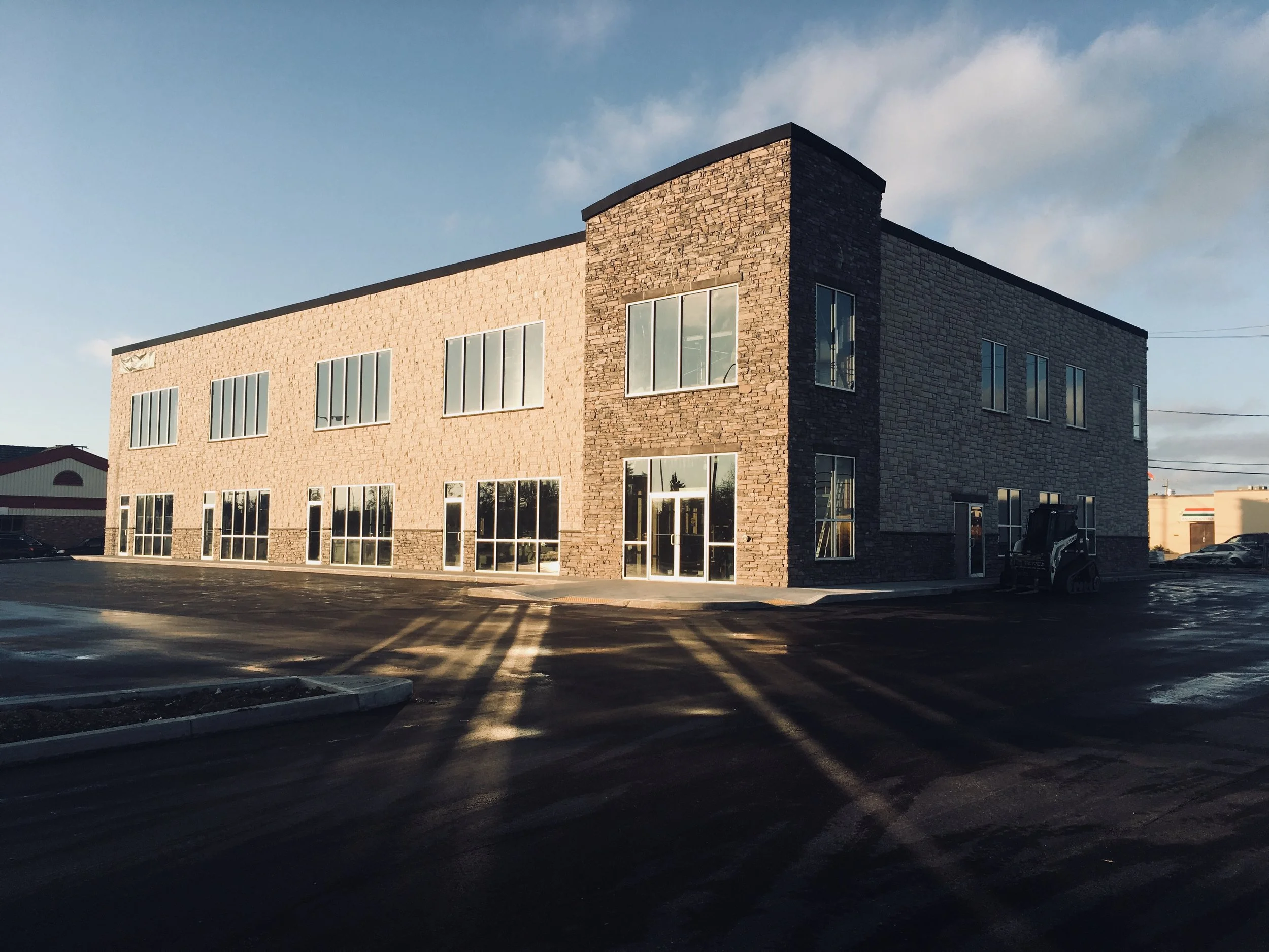 A modern multi-story brick building with large glass windows, situated in a parking lot with a few cars, under a partly cloudy sky at sunset.