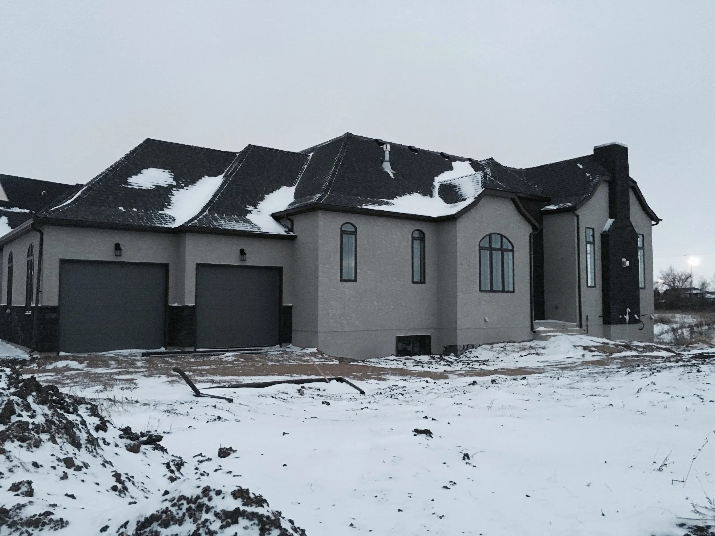 A two-story house under a cloudy sky with a dark roof partly covered in snow, large arched windows, and a chimney, with a snow-covered yard and construction debris or dirt in the foreground.