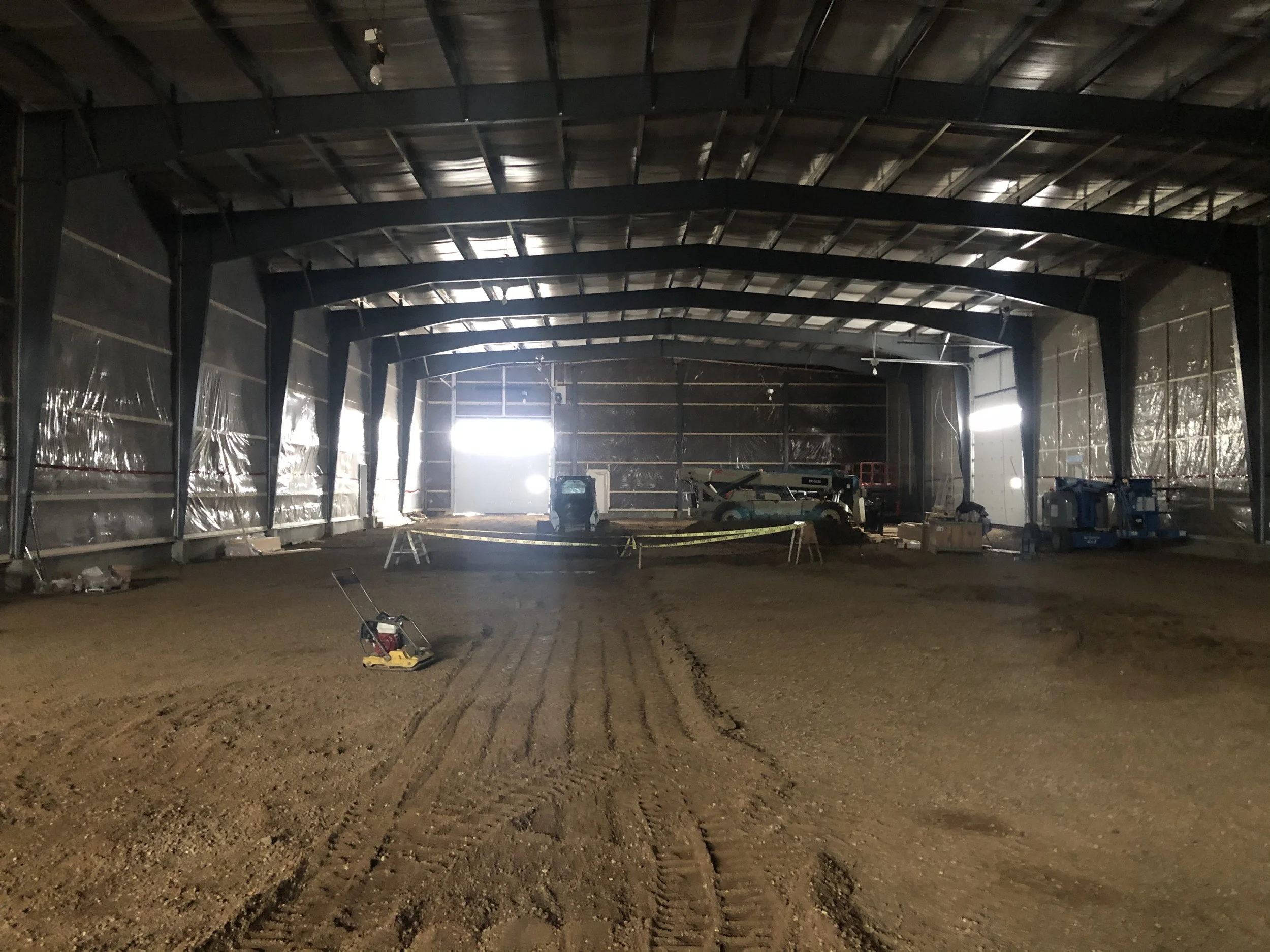Interior view of a large industrial building or warehouse under construction, with exposed metal framework and construction equipment inside.