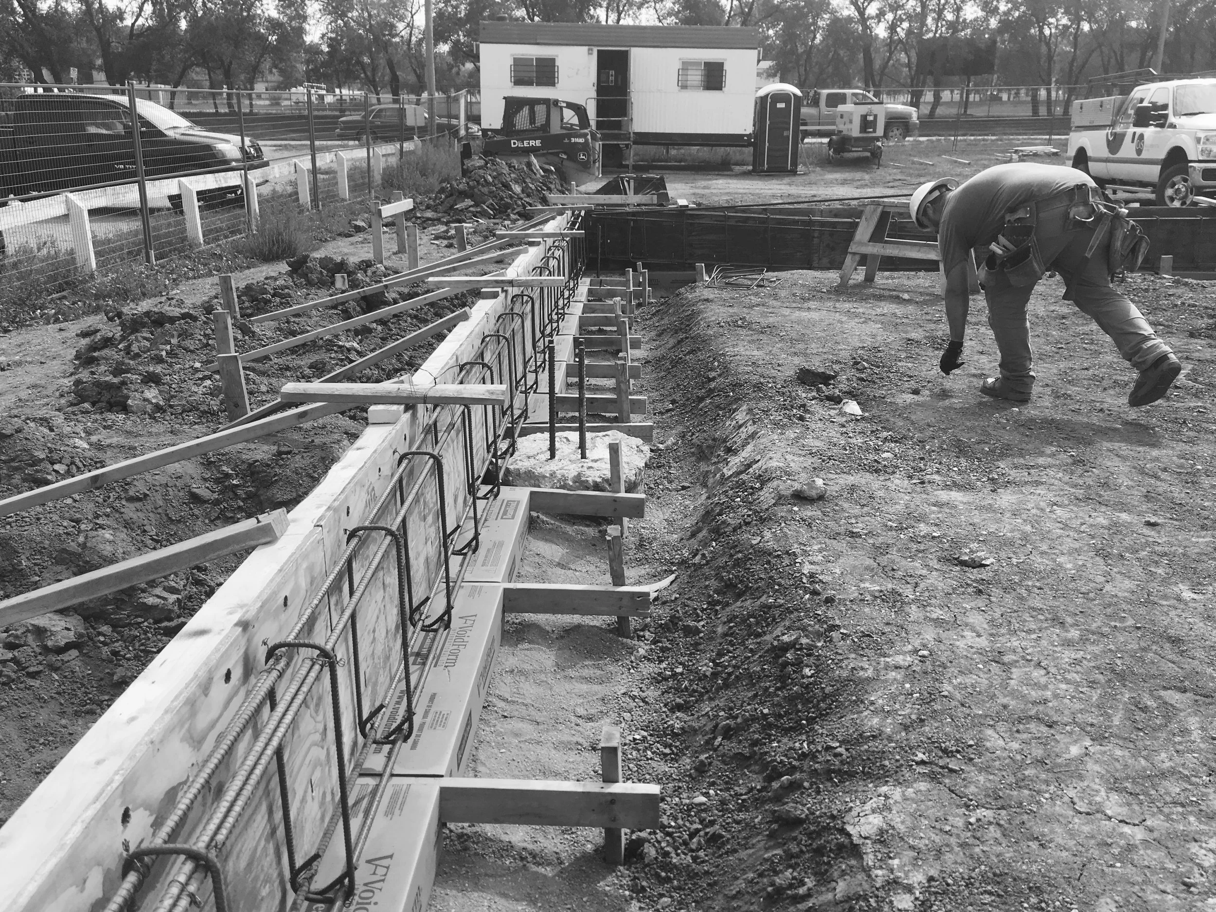 Construction worker bending over on a construction site with framing and rebar, amidst dirt and construction materials, with vehicles and a small building in the background.