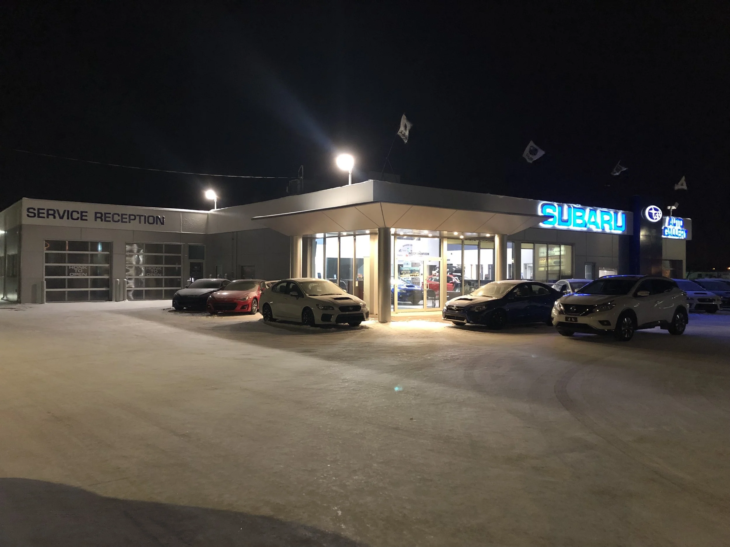 Nighttime view of Subaru dealership with illuminated neon blue sign, several cars parked outside, and service reception area, with flags waving in the dark sky.