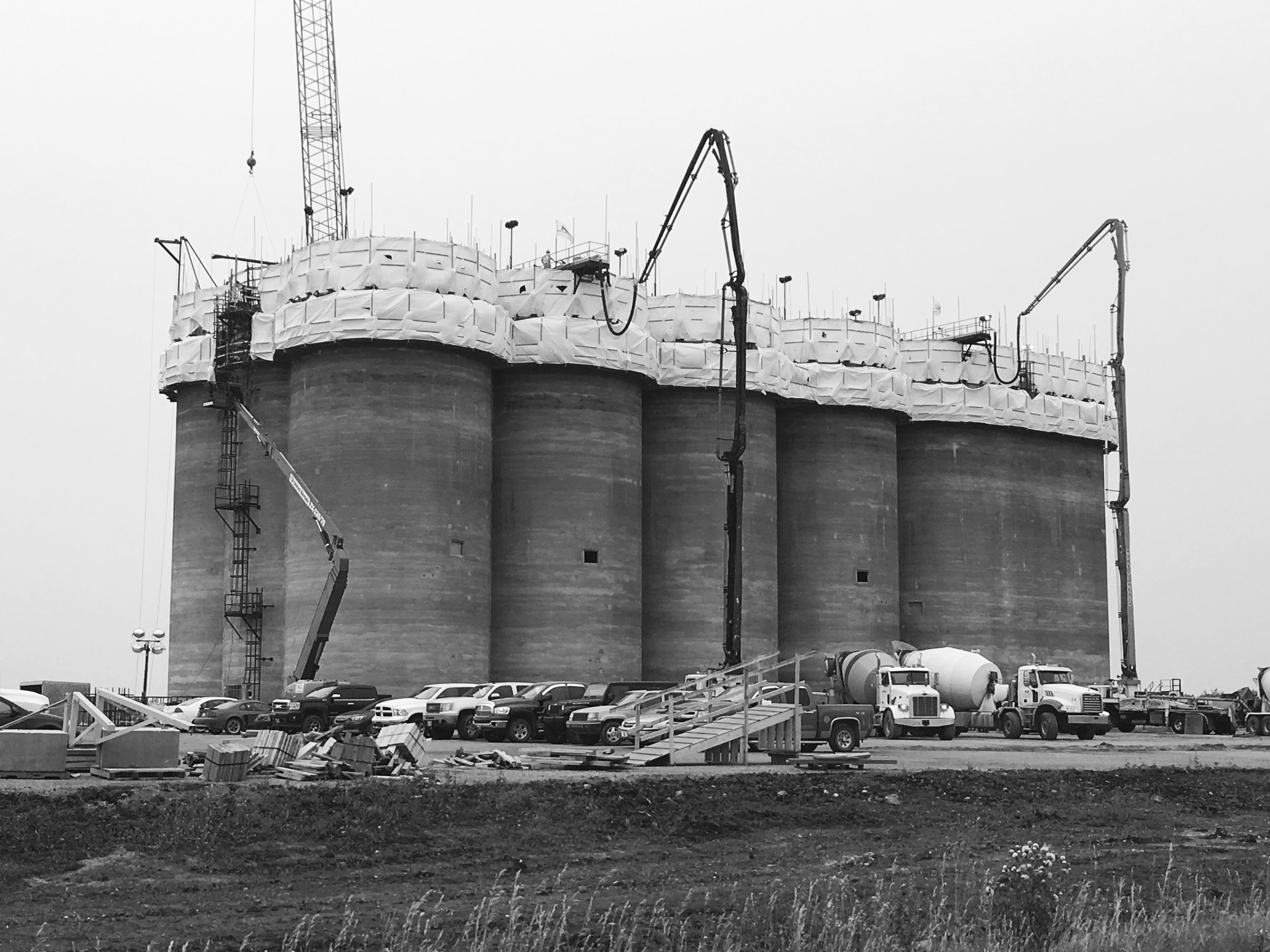 Construction site with large industrial tanks, cranes, and construction equipment. Cars parked in front of the site.