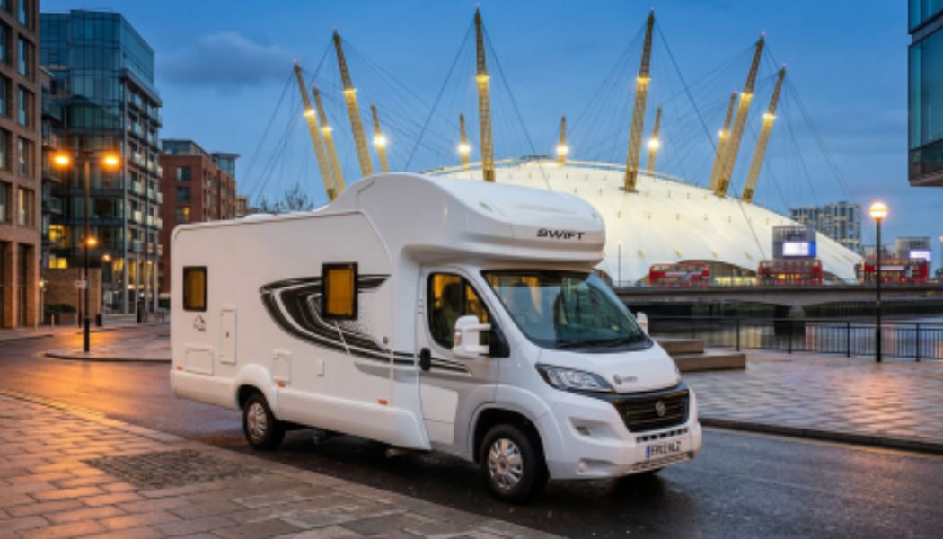 Large white Swift motorhome parked on a paved city street in London at twilight with the illuminated yellow spikes of the O2 Arena dome visible in the background.