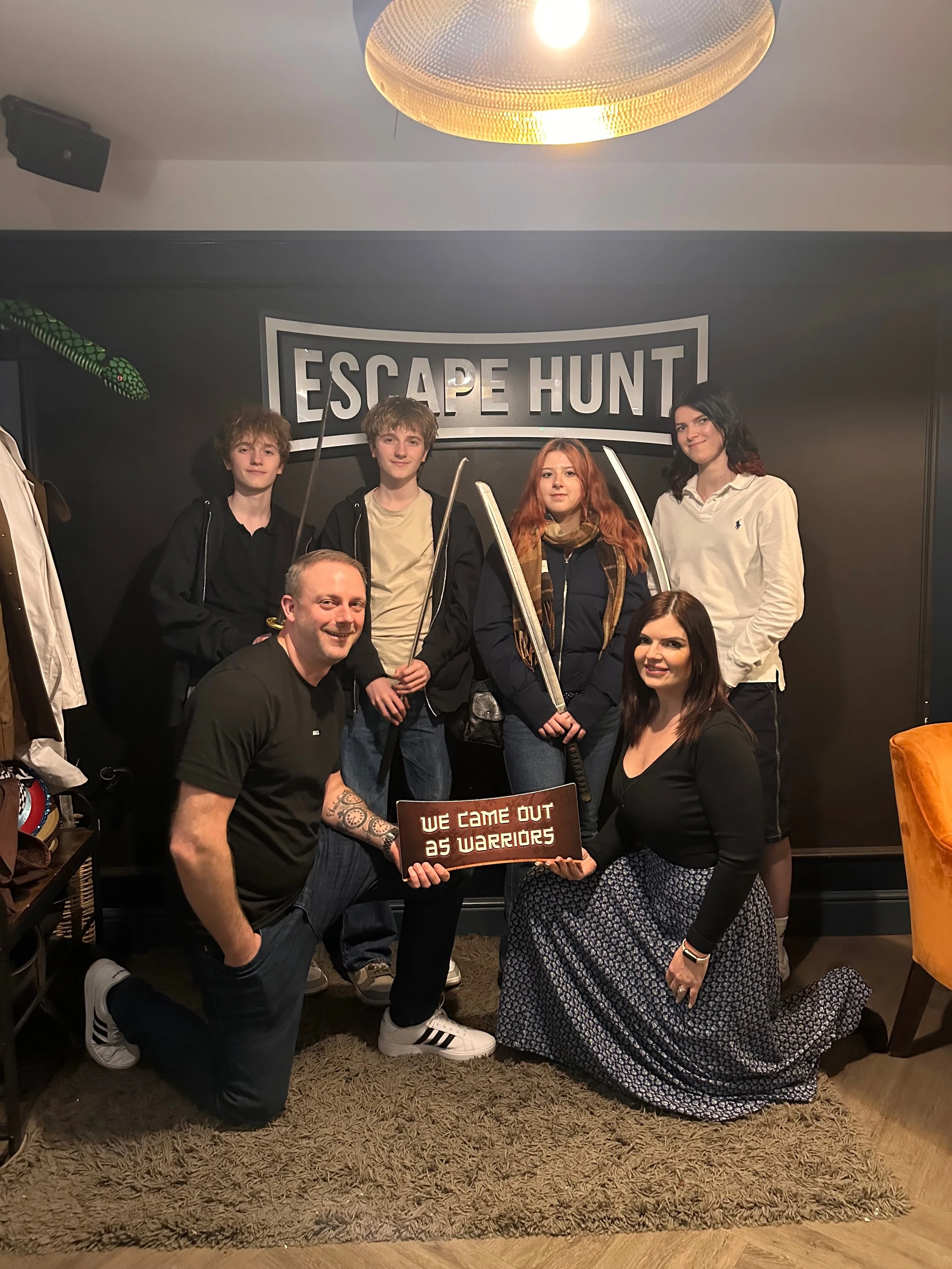 A group of seven people posing in front of an "Escape Hunt" sign indoors. They are holding toy swords and one person is holding a sign that says "We Came Out as Warriors." Everyone is smiling or has a neutral expression.