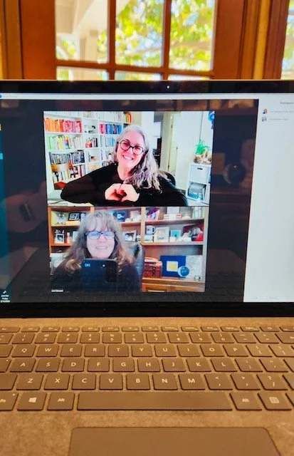 Two women are participating in a video call on a laptop. The woman on the top has gray hair, glasses, and is smiling with her hands folded; she is in a room with bookshelves. The woman on the bottom has light brown hair, glasses, and is also smiling; she is in a room with bookshelves and various objects. The laptop is on a table or desk near a window with visible foliage outside.