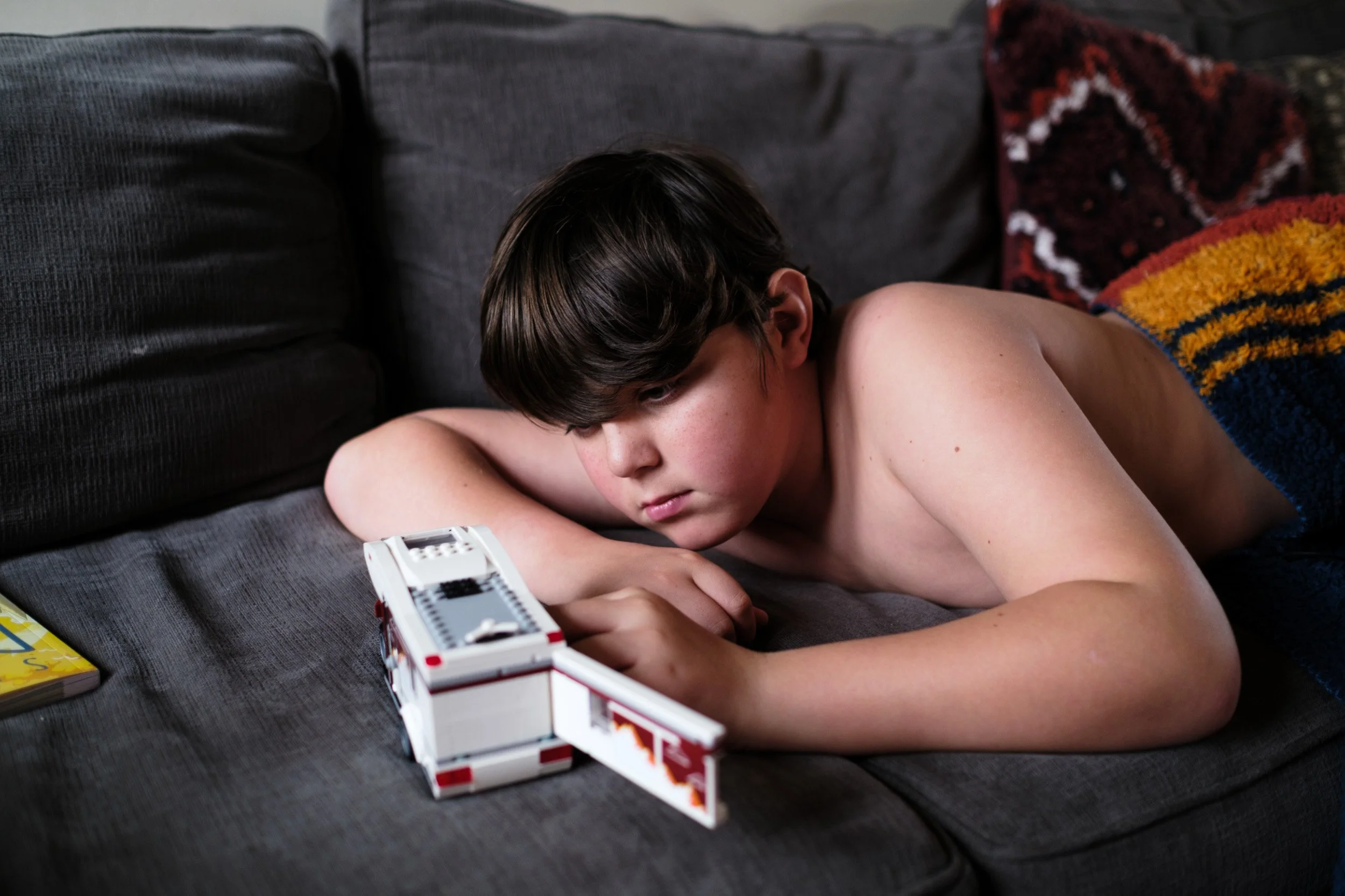 Boy rests his head on his arm on a sofa, watching a small toy in front of him