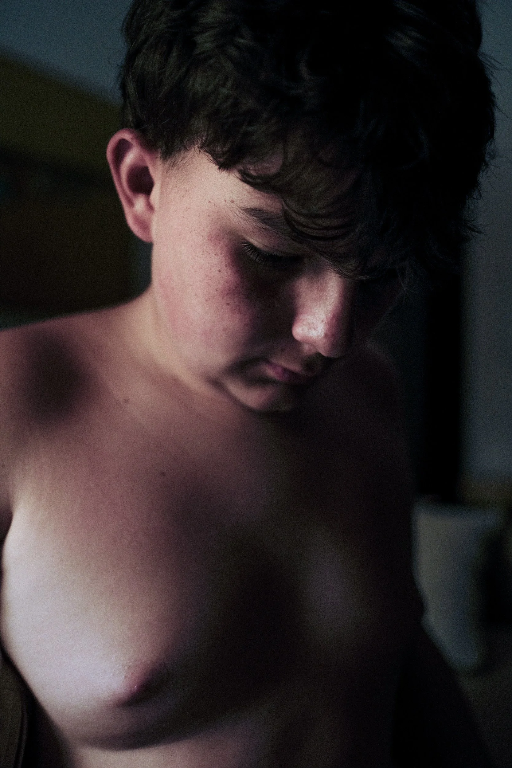 Boy looks down in a dark room, his face and shoulder caught in a sliver of light