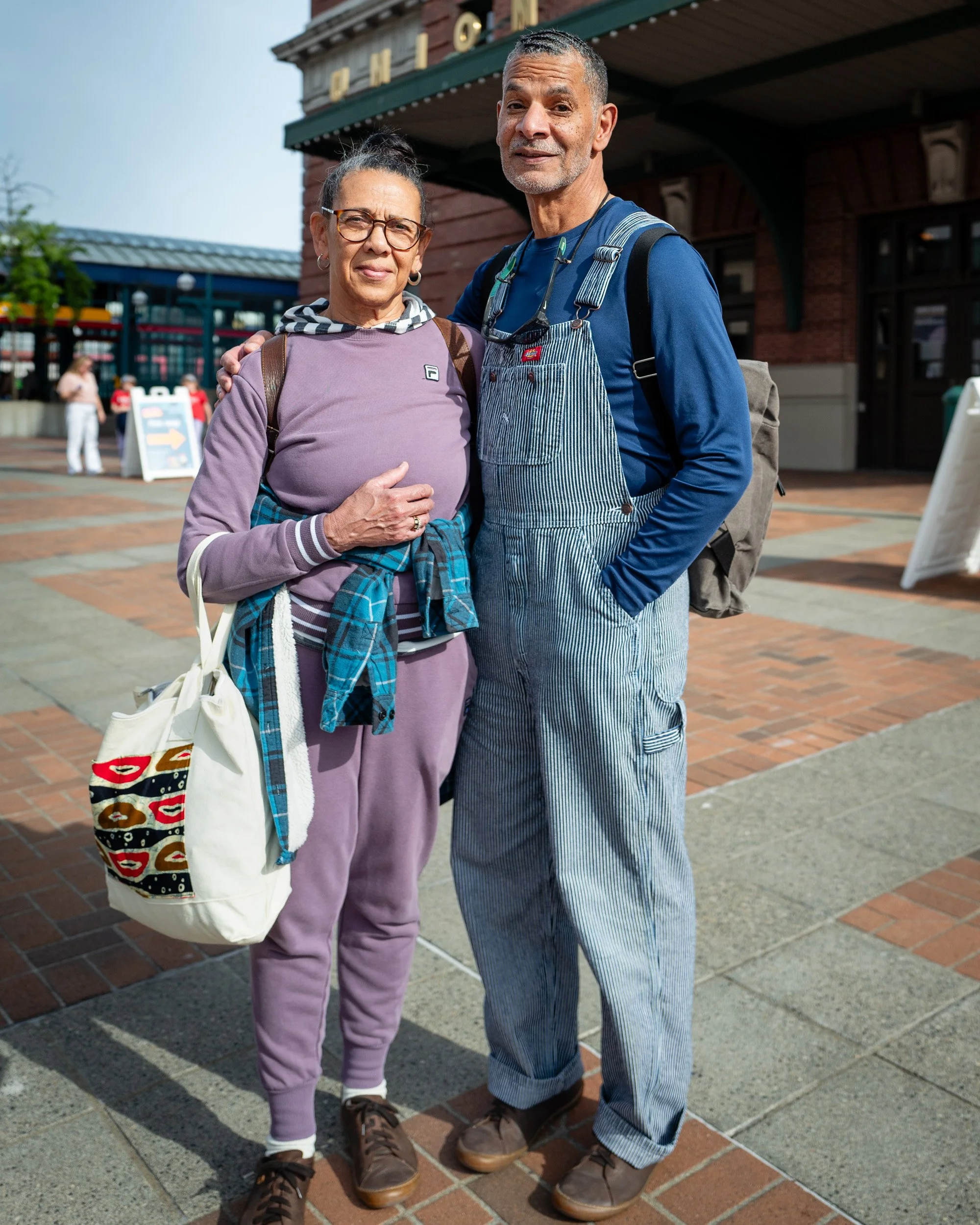 Older couple, she in lavender and he in denim overalls, stand side by side on a brick plaza