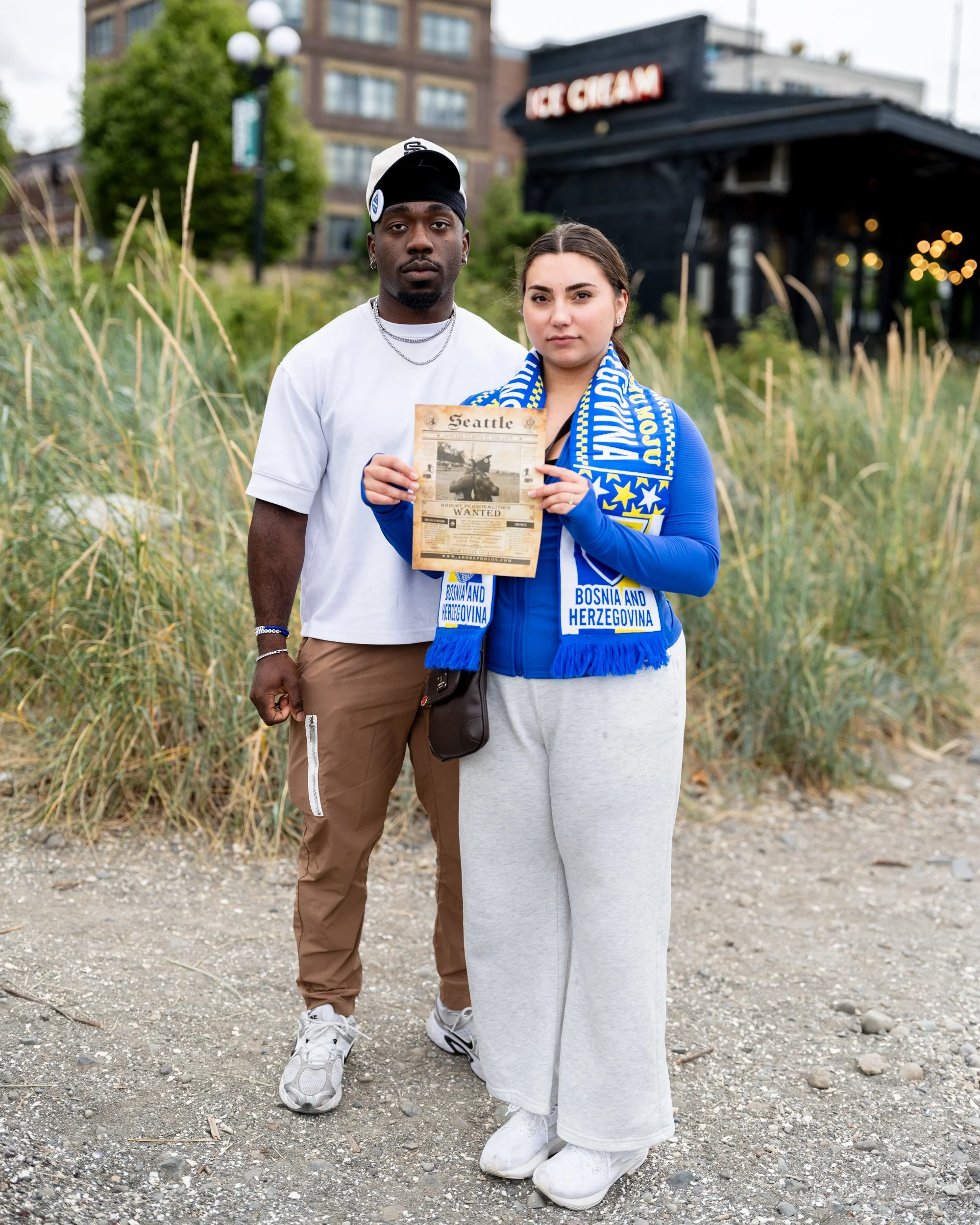 Young man and woman in a blue soccer scarf stand together holding a printed flyer beside tall grasses