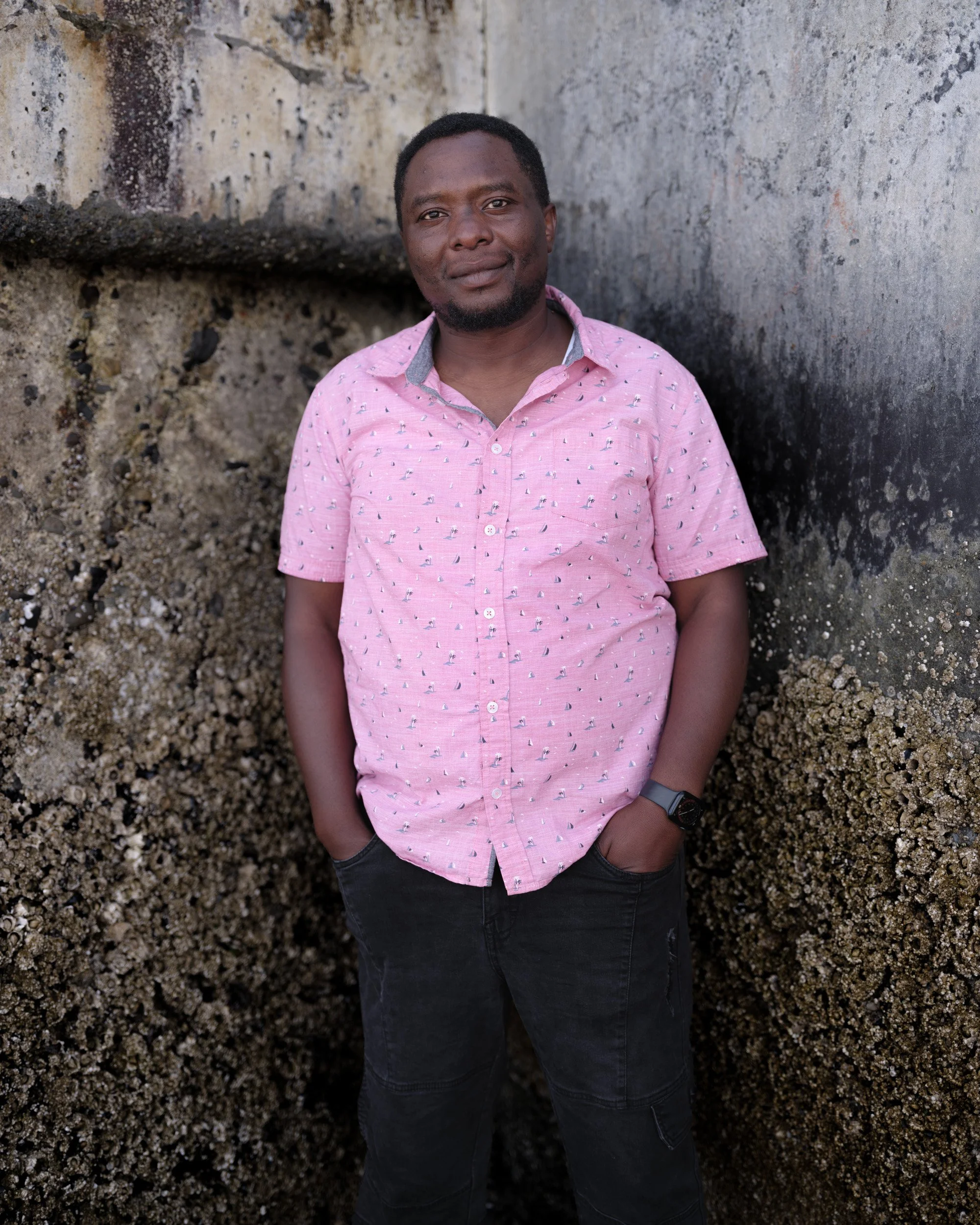 Man in a pink short-sleeved shirt stands against a weathered concrete wall