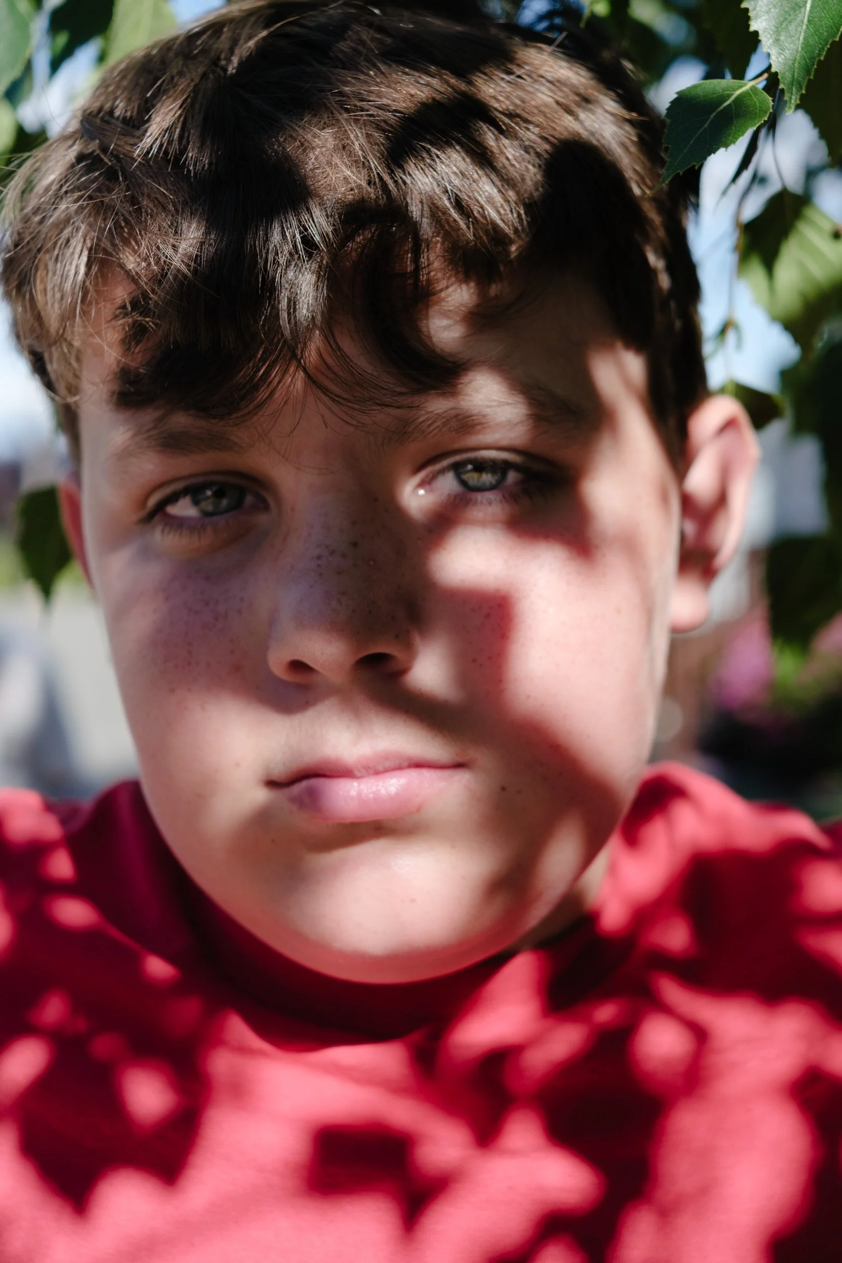 Close-up portrait of a boy in a red shirt, his face dappled with leaf shadows