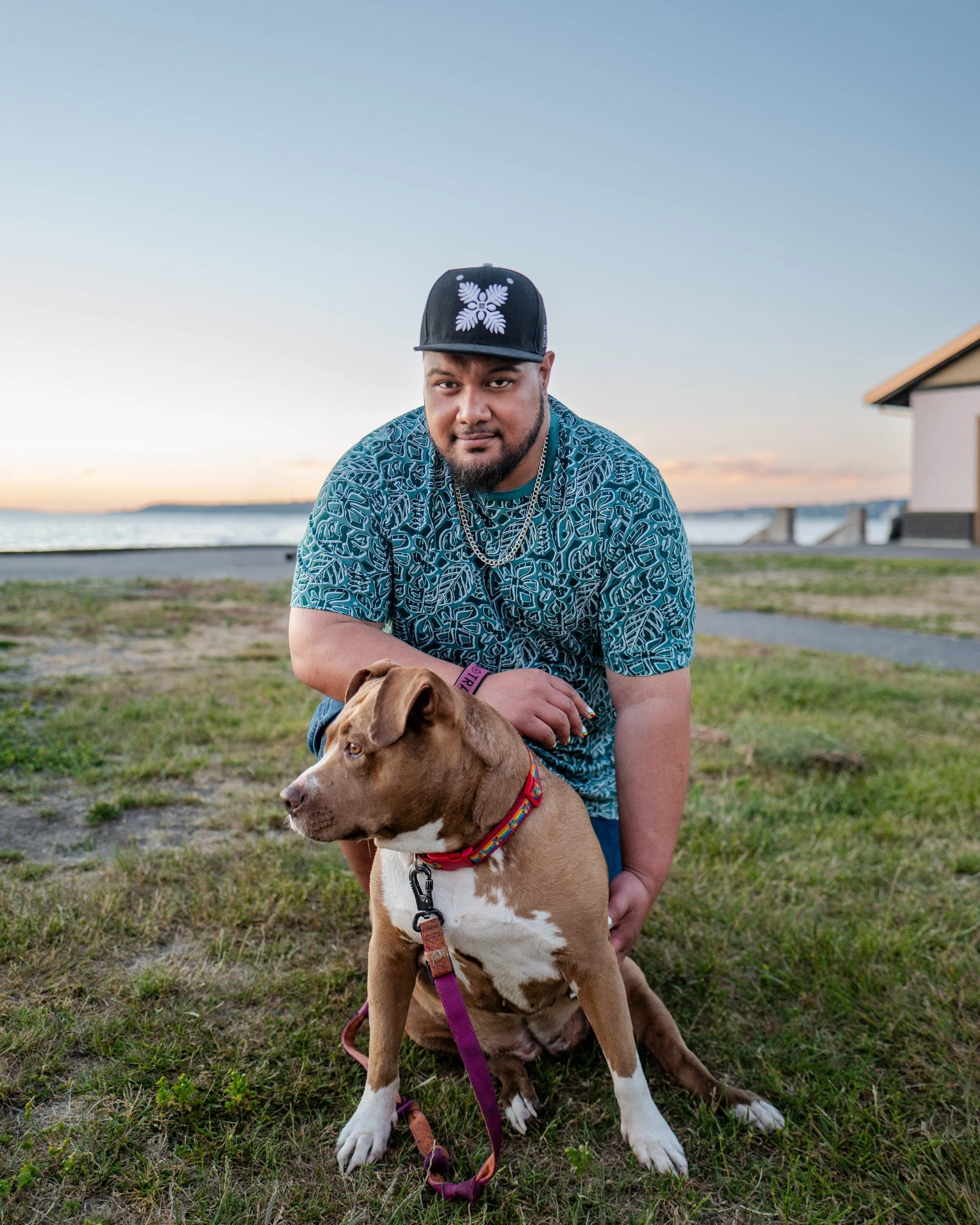 Man in a baseball cap crouches with a brown and white dog on a grassy waterfront at sunset