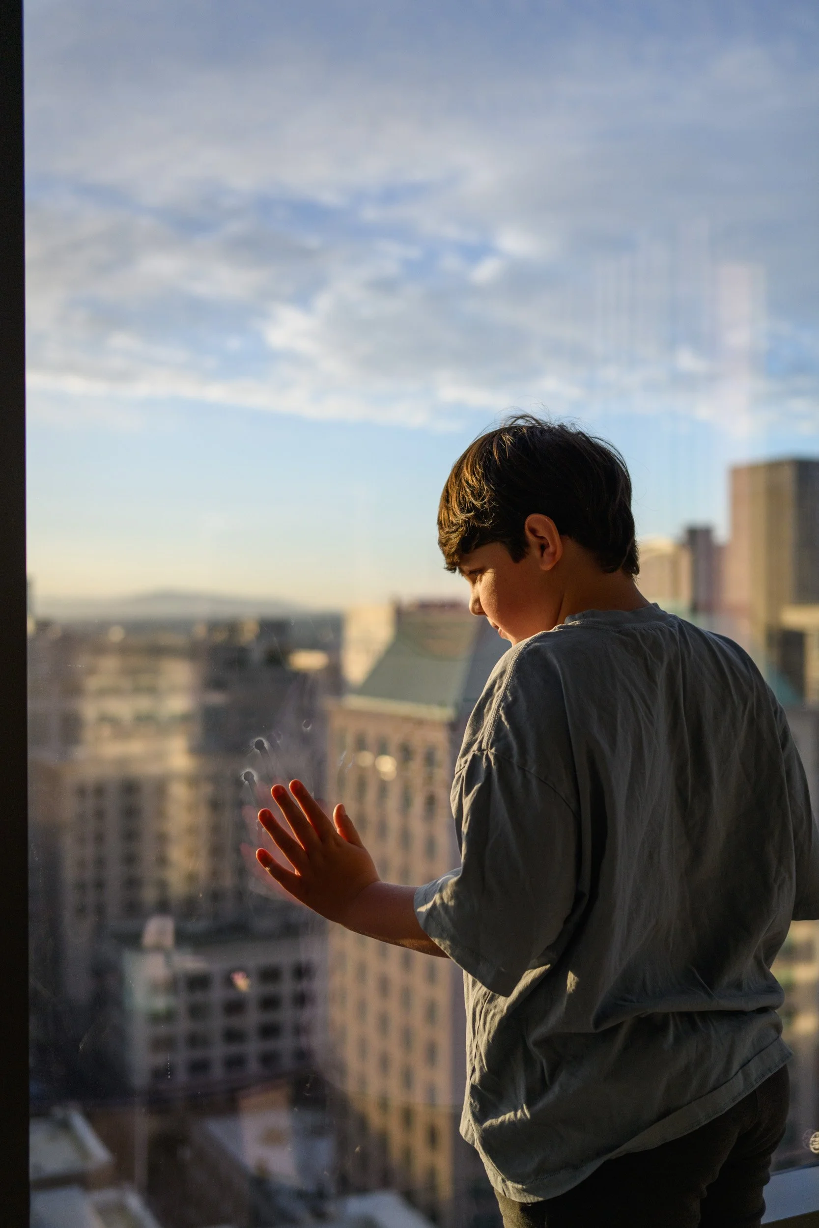Boy touches a high-rise window overlooking a city skyline in late-day light