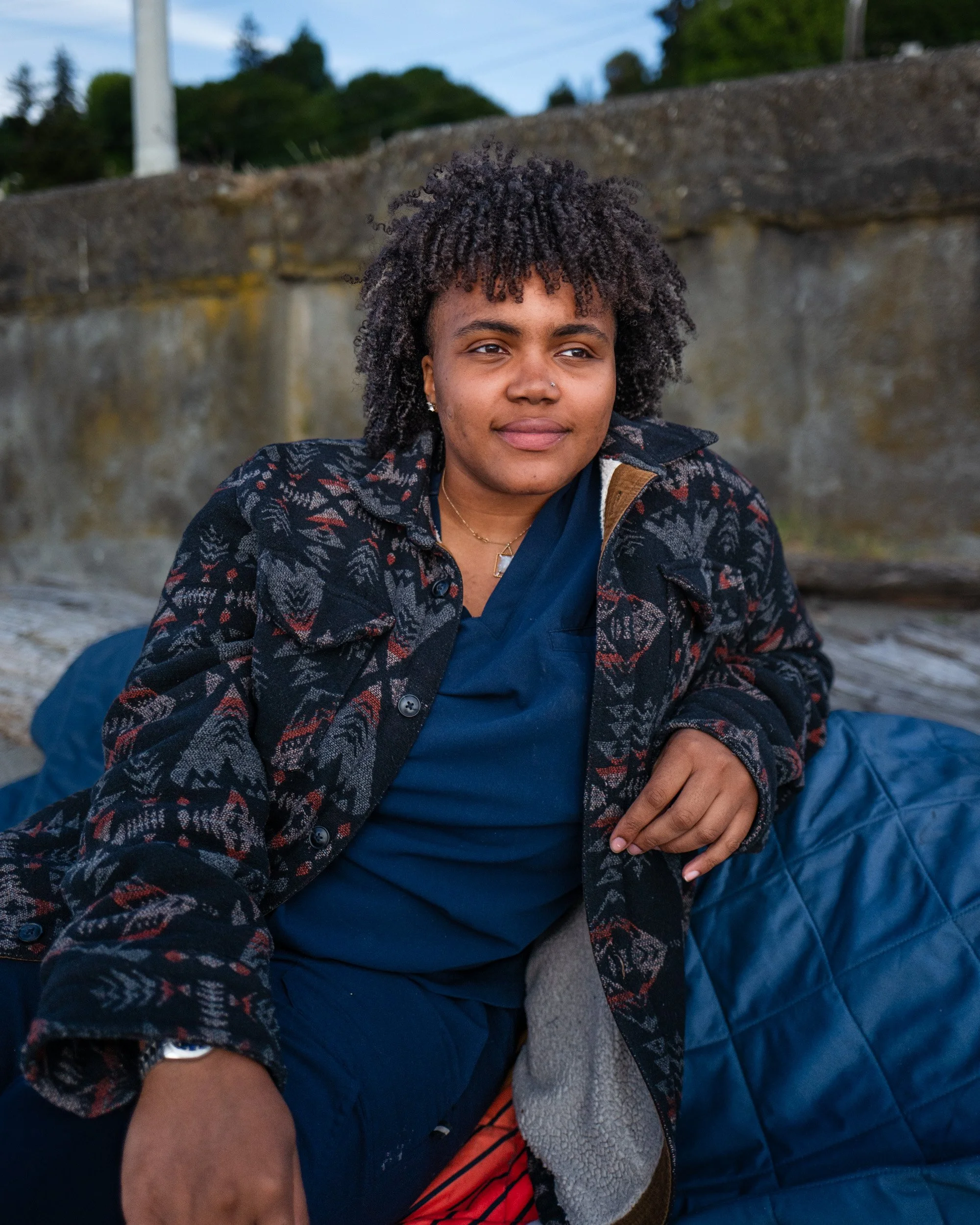 Woman with curly hair sits against a stone wall wrapped in a patterned jacket and blue blanket