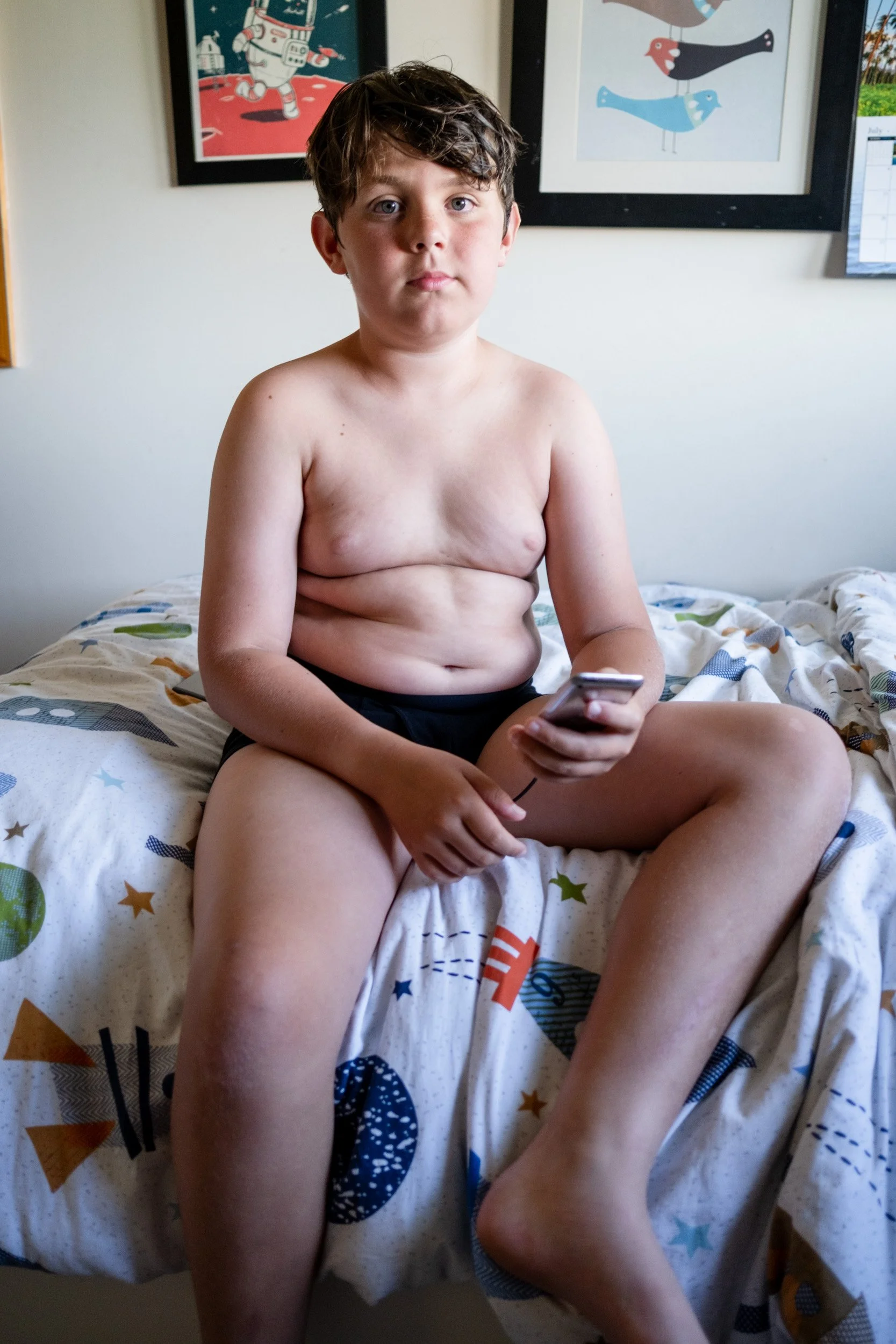 Boy sits on a bed with a rocket-print duvet, holding a phone and looking at the camera