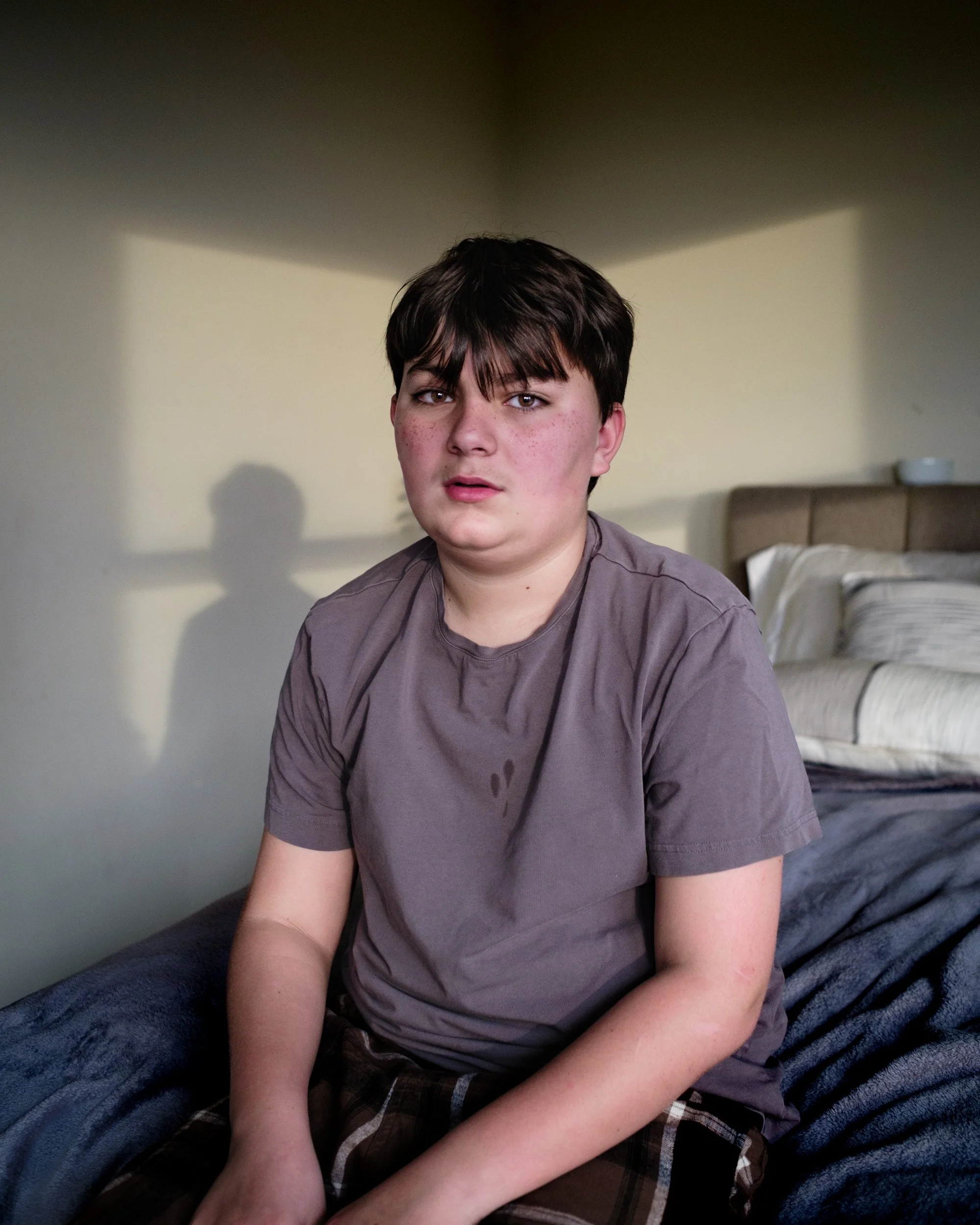 Boy in a gray T-shirt sits on a bed in warm window light, his shadow cast on the wall behind him
