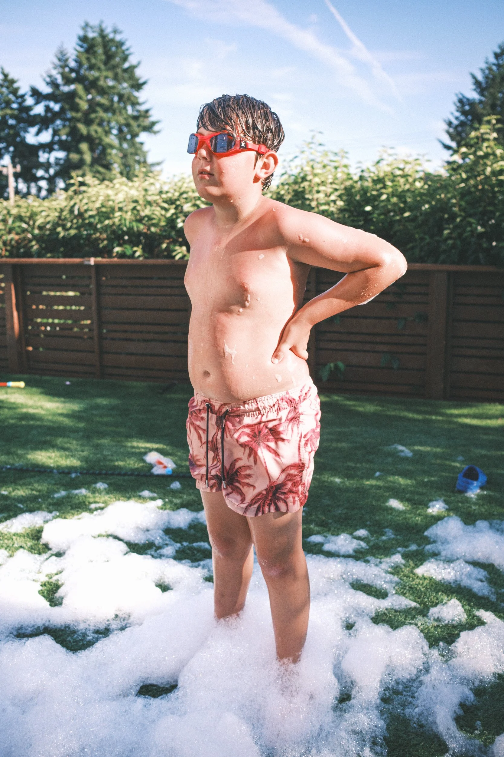 Boy in red sunglasses and swim trunks stands with hands on hips in soap foam on a backyard lawn