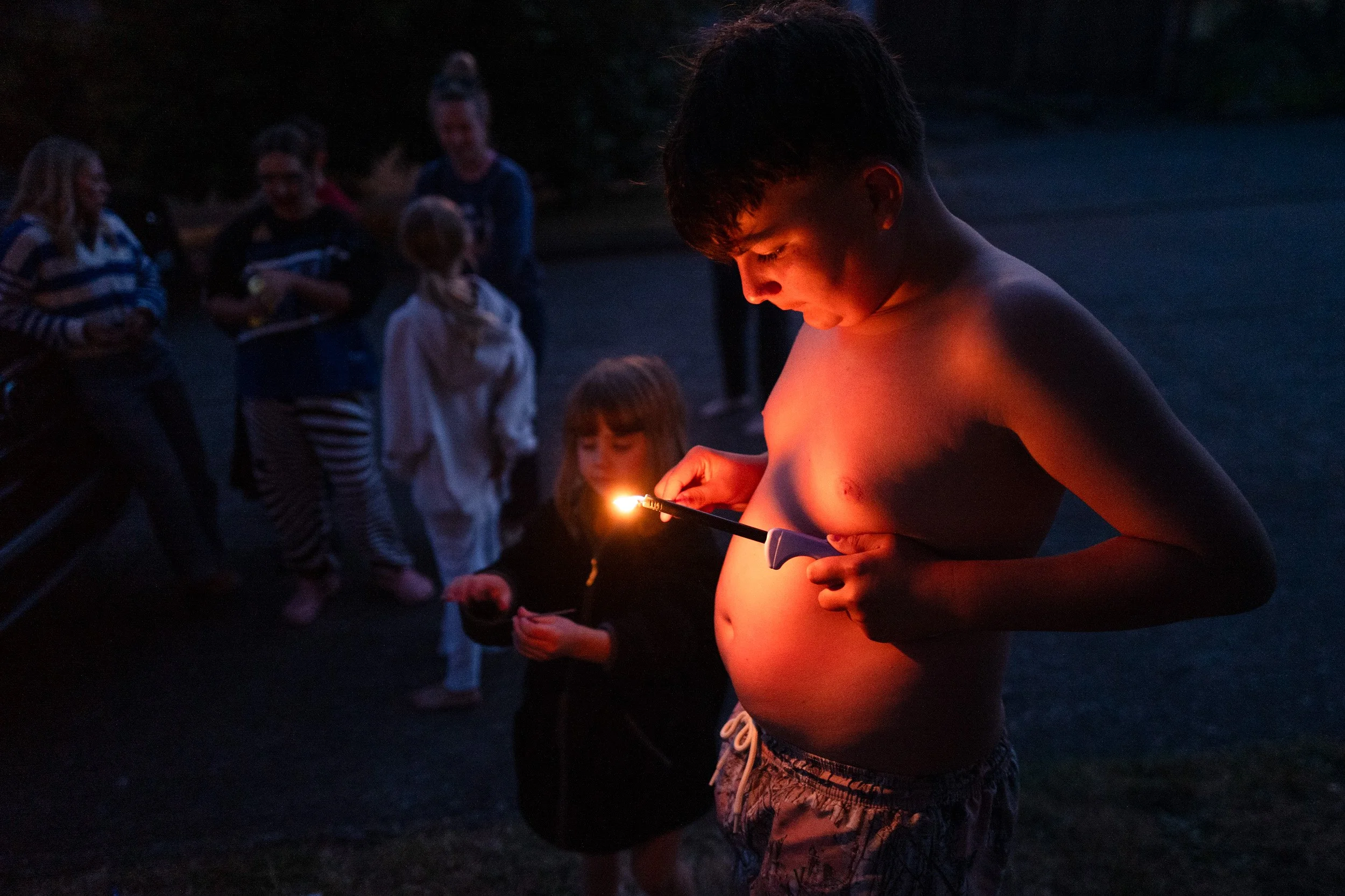 At dusk, a small flame lights a boy's face and chest as children gather in a dark backyard