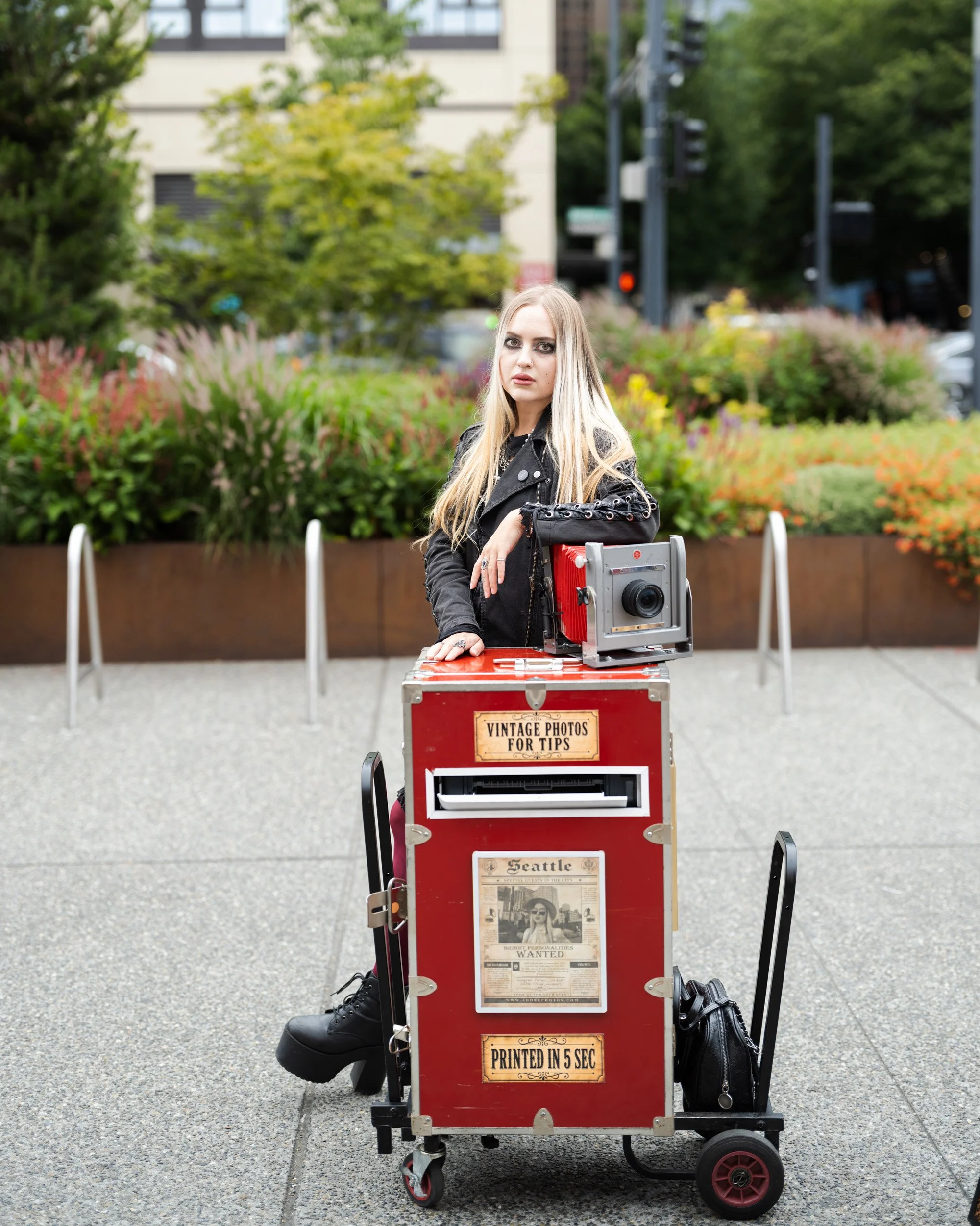 Young woman with long blond hair stands beside a red vintage cart topped with a camera on a city plaza