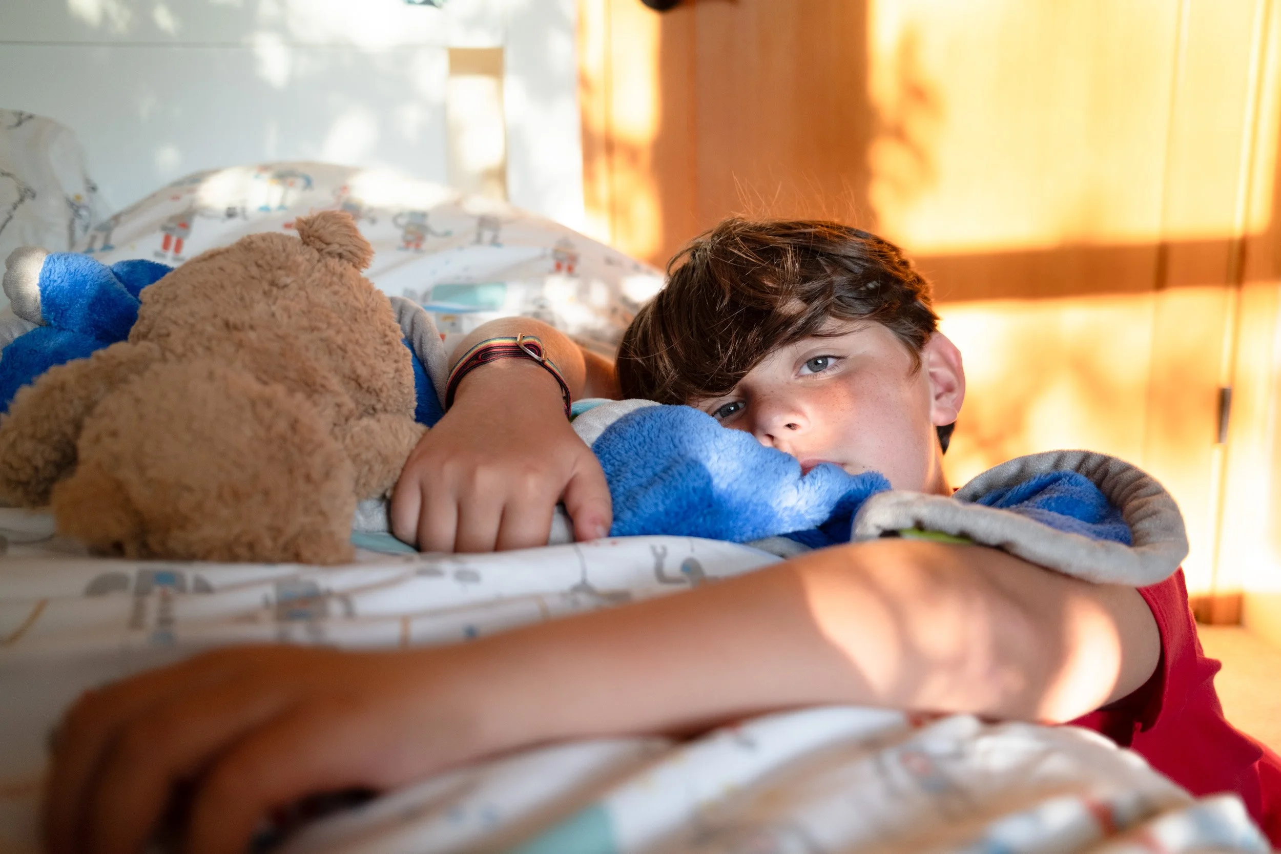Boy lies in bed beside a teddy bear, morning sunlight falling across the room