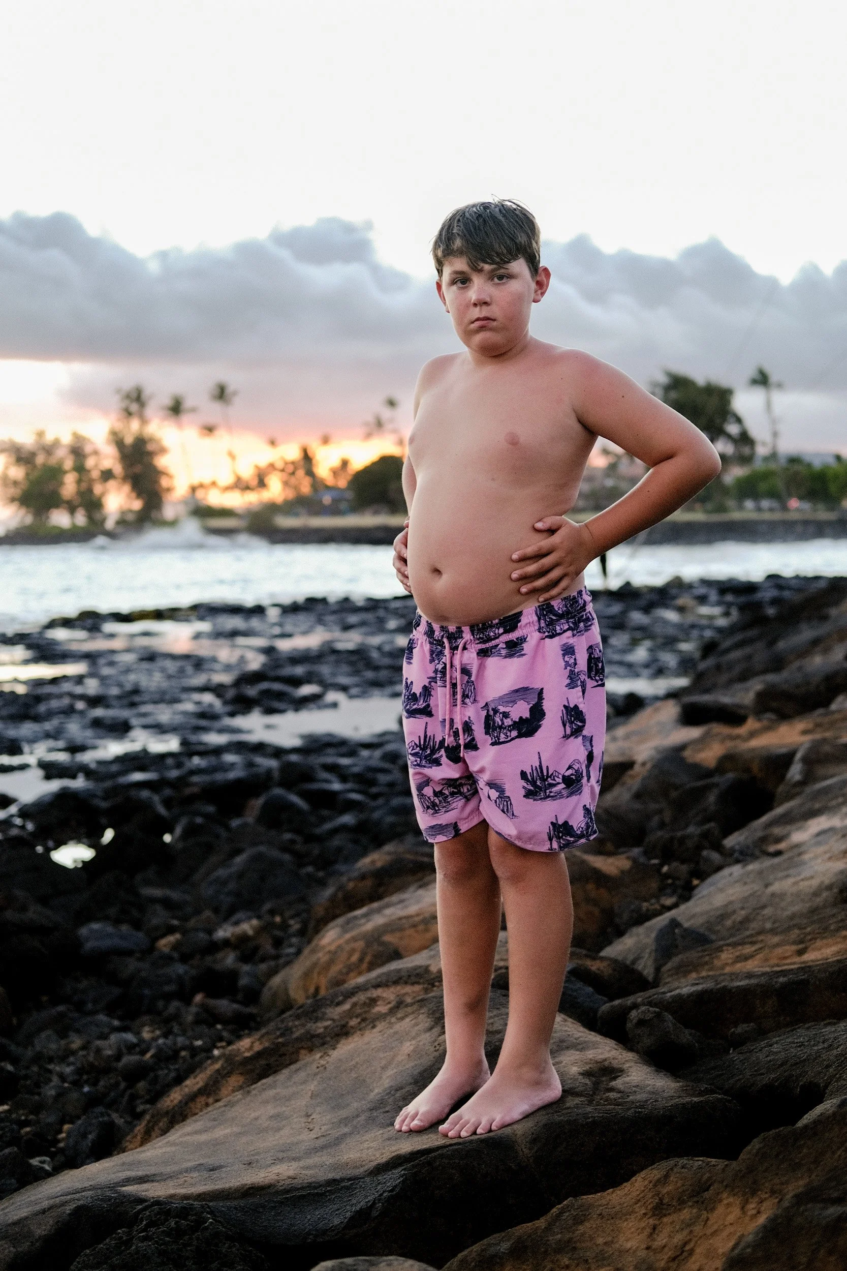 Boy in purple swim trunks stands on dark volcanic rocks at the ocean's edge at sunset