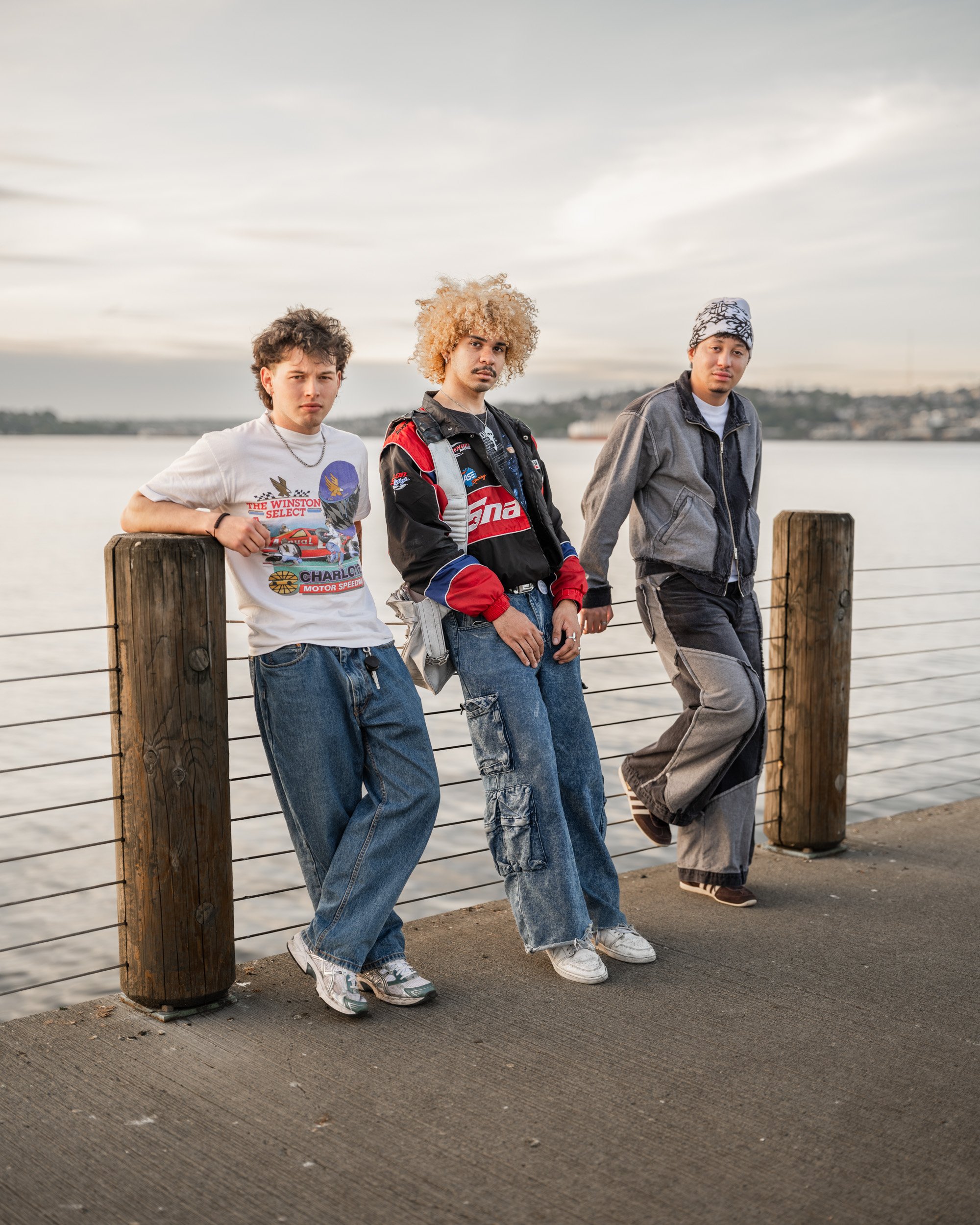 Three young people lean against wooden posts on a Seattle waterfront under an overcast sky