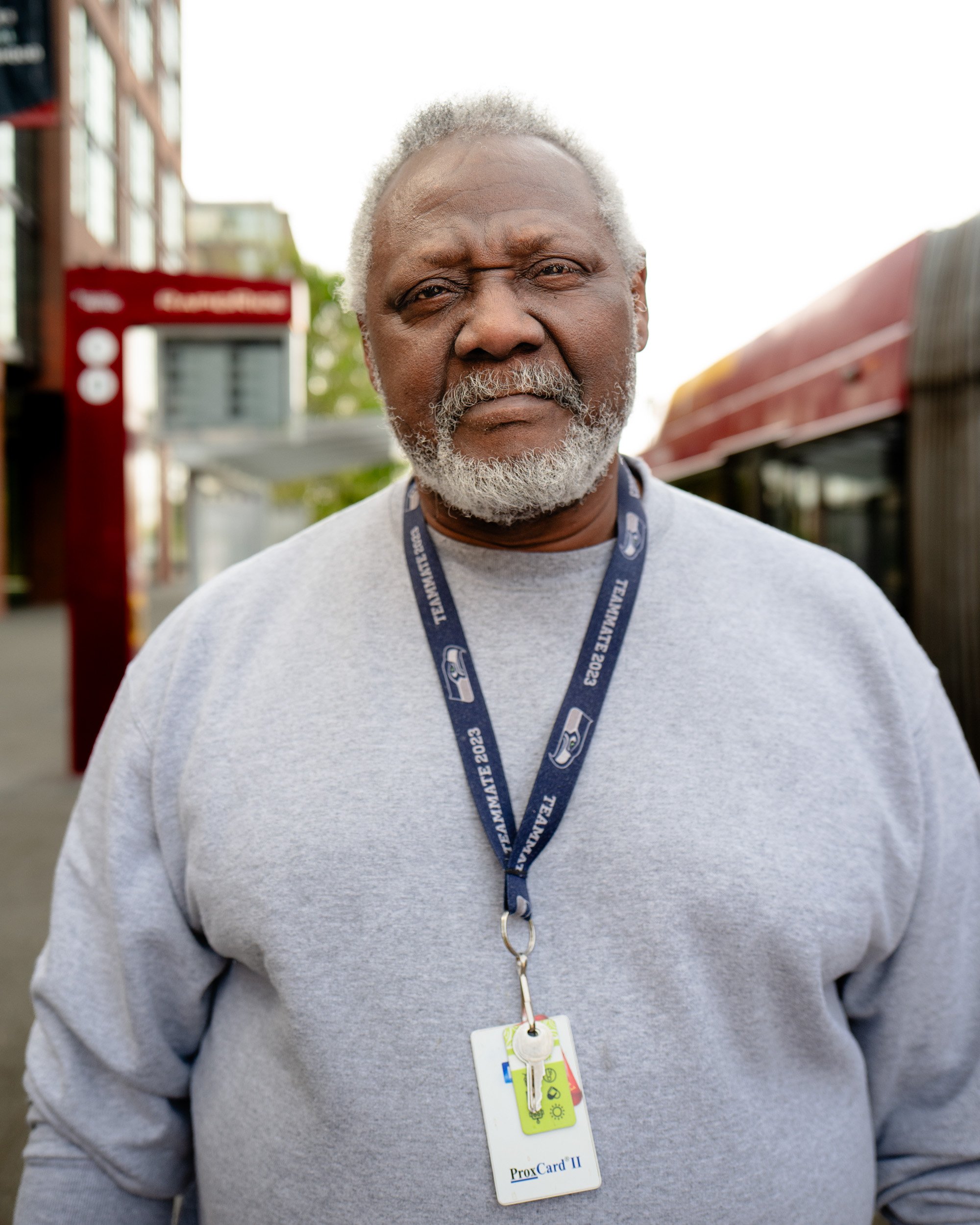 Older man with a white beard and a lanyard badge stands on a city sidewalk