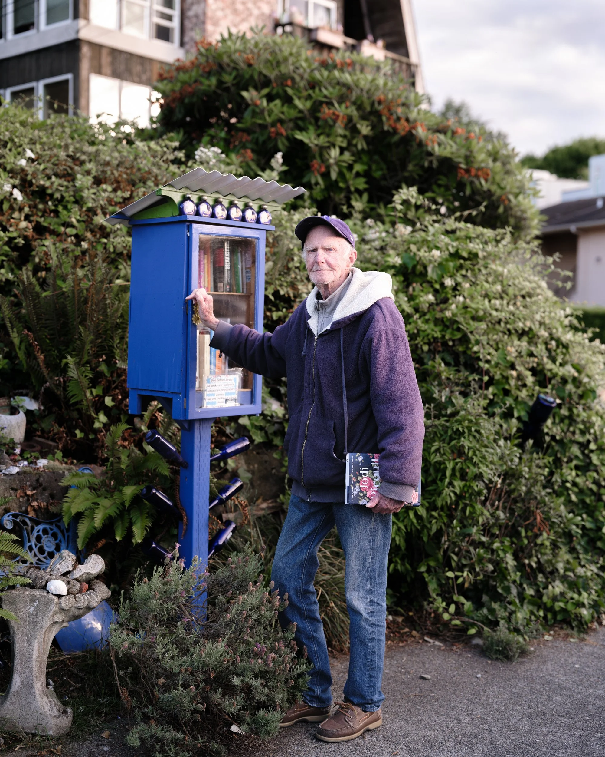 Older man in a cap stands beside a blue Little Free Library surrounded by garden shrubs