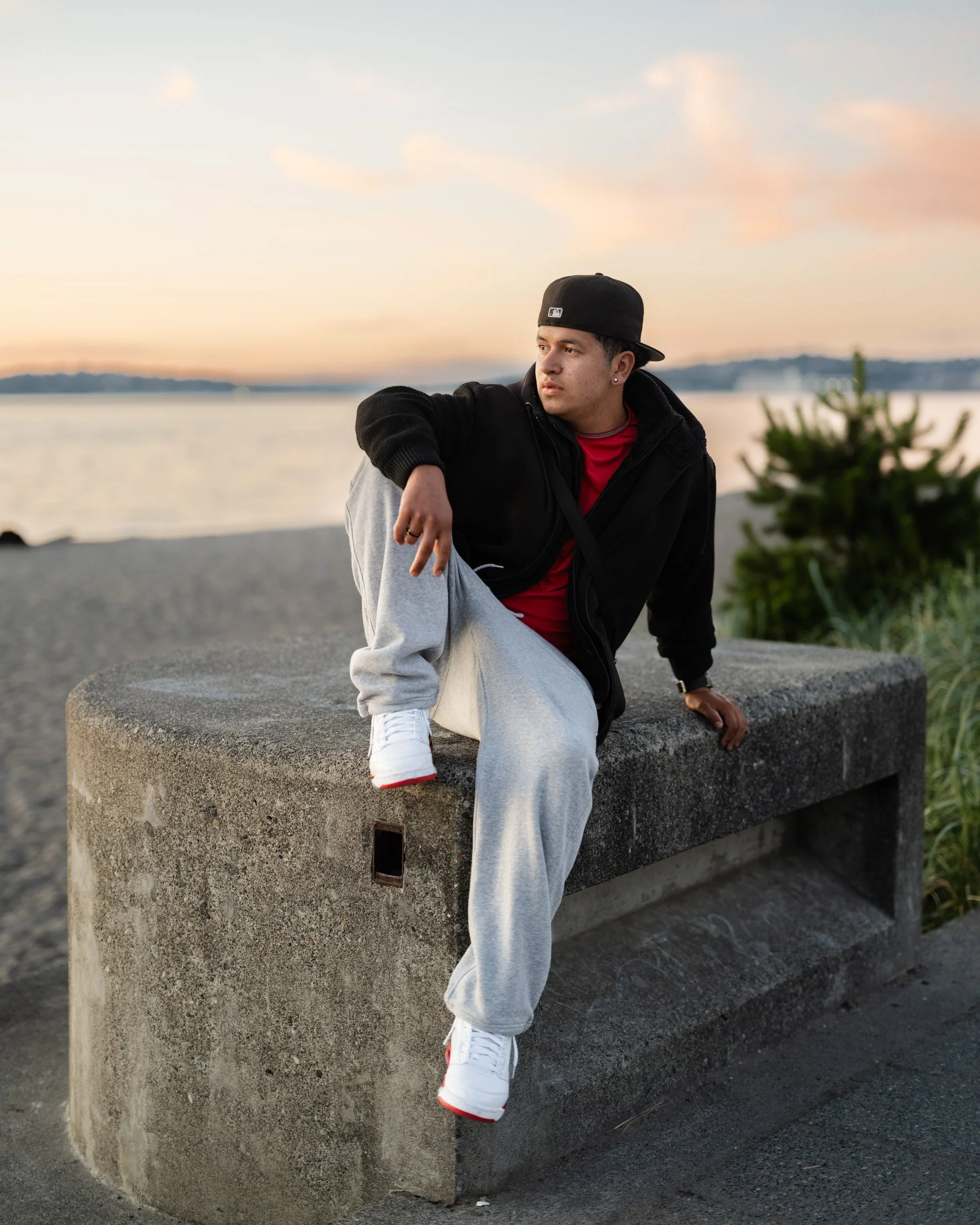 Young man in a backwards cap sits on a concrete wall by the water at sunset