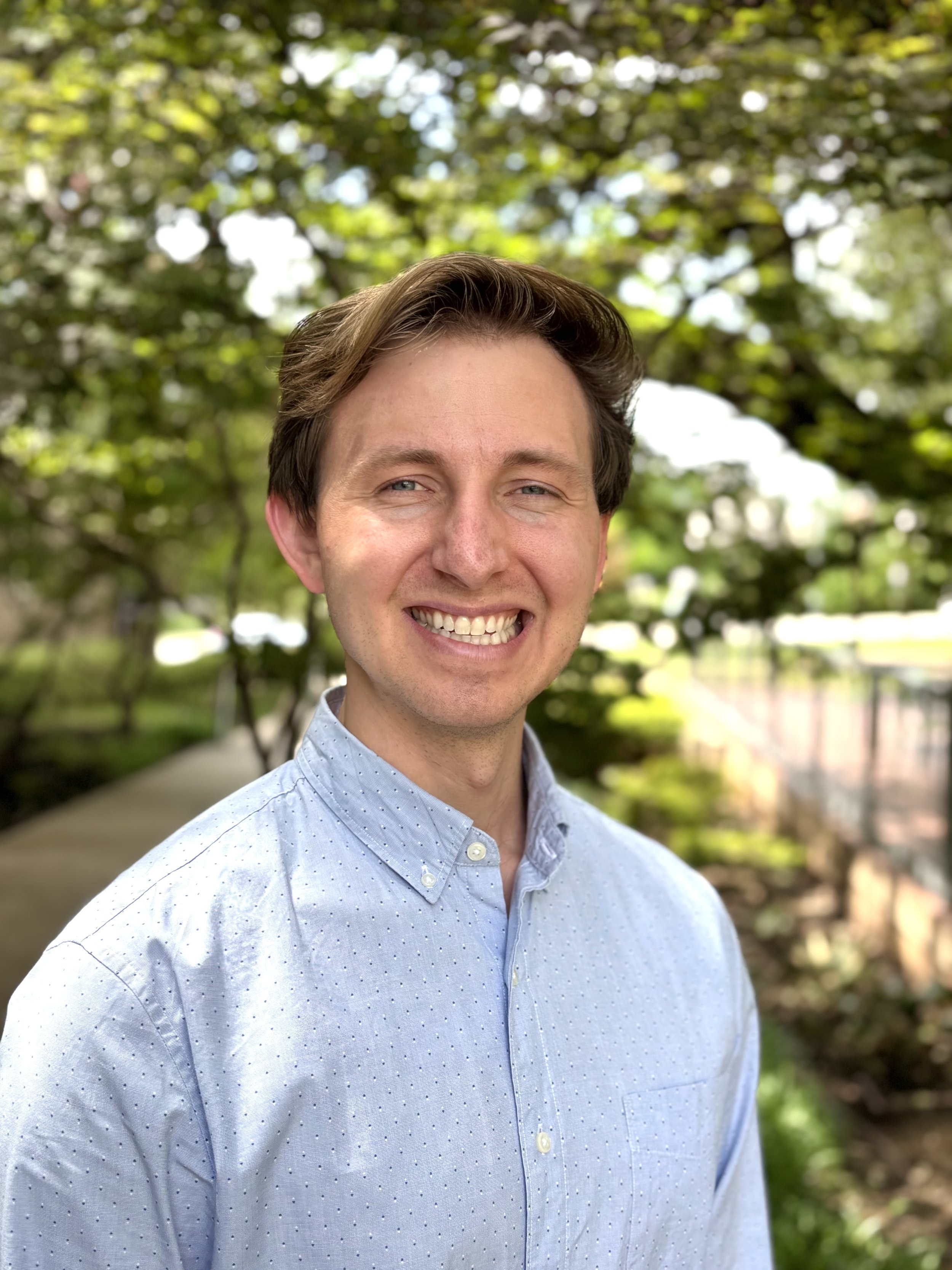 A smiling man with light brown hair and blue eyes, wearing a light blue, polka-dotted button-up shirt, standing outdoors with a background of green trees and sunlight.