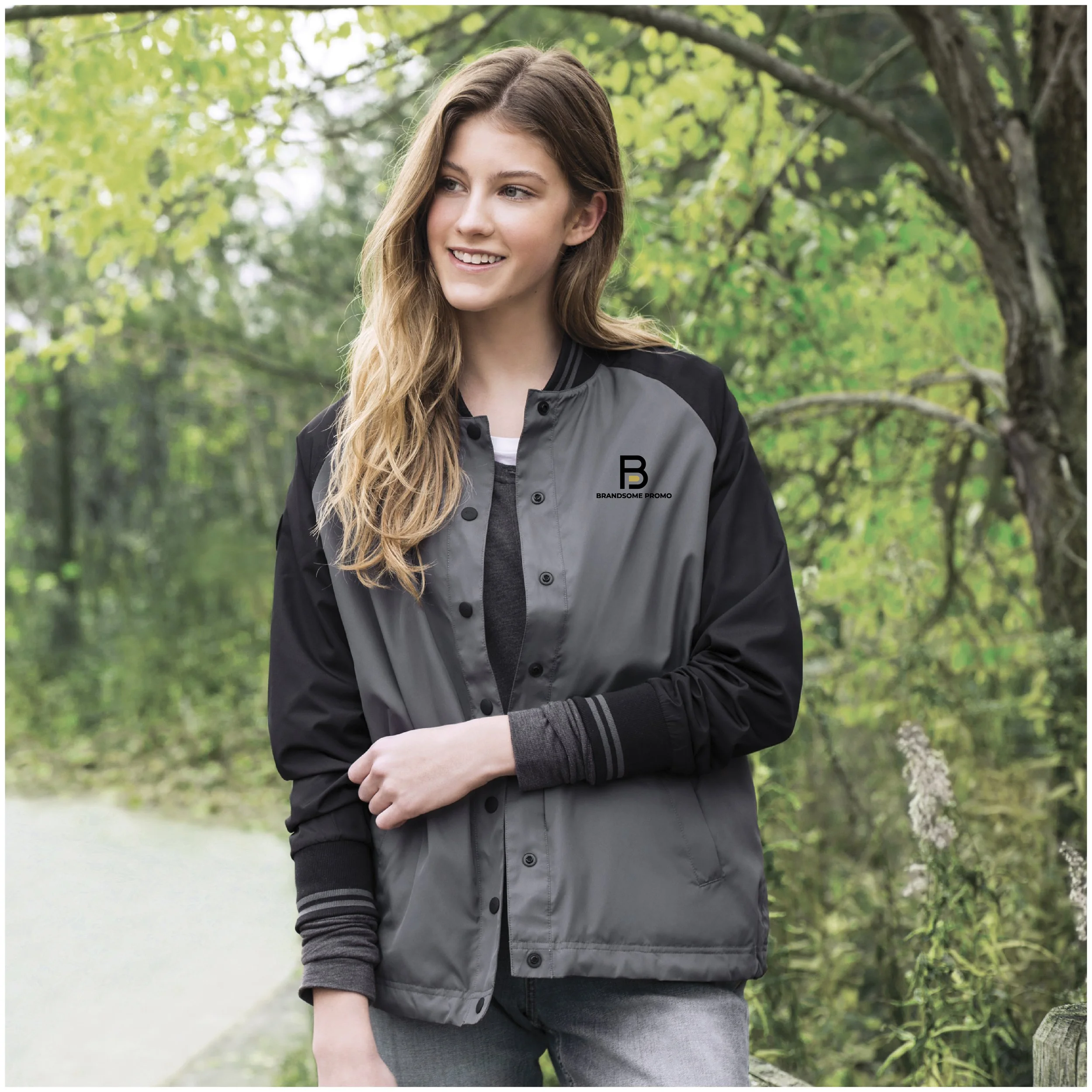 A young woman with long wavy hair smiling outdoors in a park, wearing a gray and black jacket with a logo on the chest, with green trees in the background.