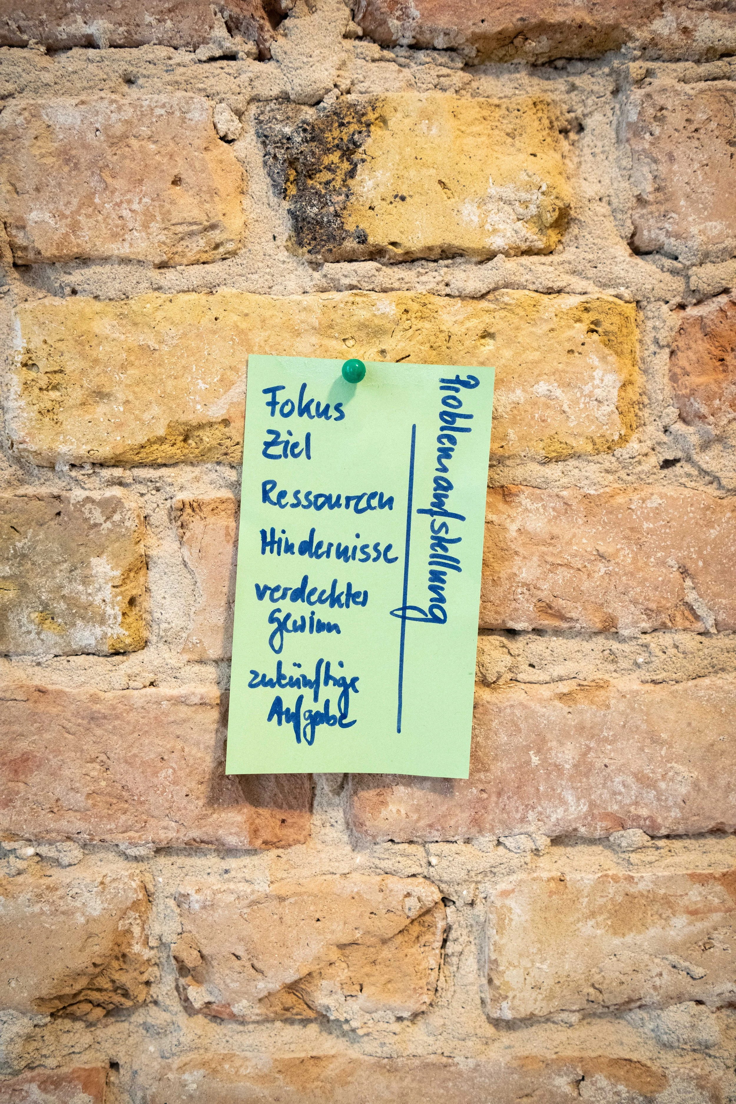 Ein gelber Zettel mit handbeschriebenem Text hängt an einer braunen Backsteinmauer. Der Text enthält Stichworte wie 'Fokus', 'Ziel', 'Ressourcen', 'Hindernisse', 'verdeckte', 'äußere' sowie eine Linie und die Website 'bauausshandwerk'.
