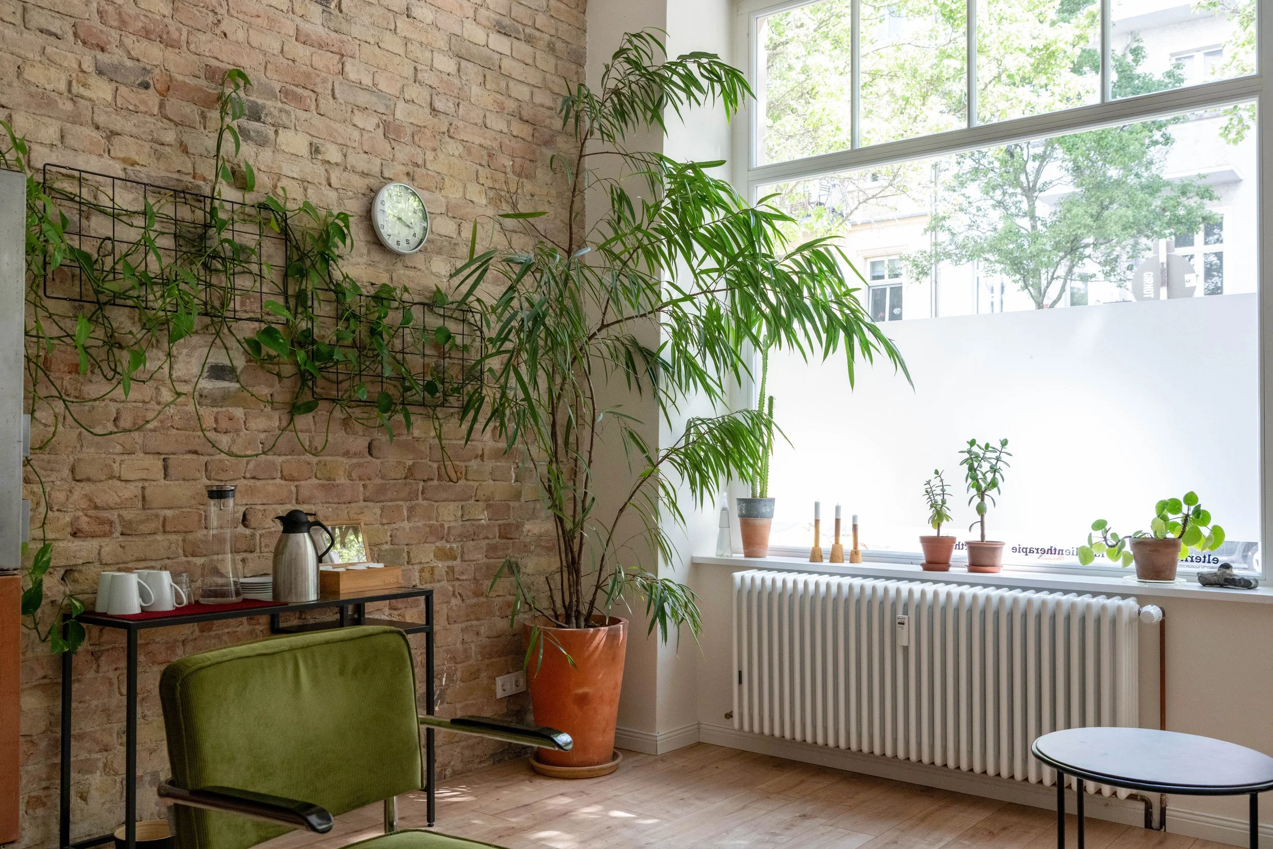 Innenraum mit Backsteinwand, großen Fenster, mehreren Pflanzen in Töpfen, einer Uhr an der Wand, einem Sessel, einem Tisch mit Kaffeetassen, einem Wasserkocher und weiteren Gegenständen.