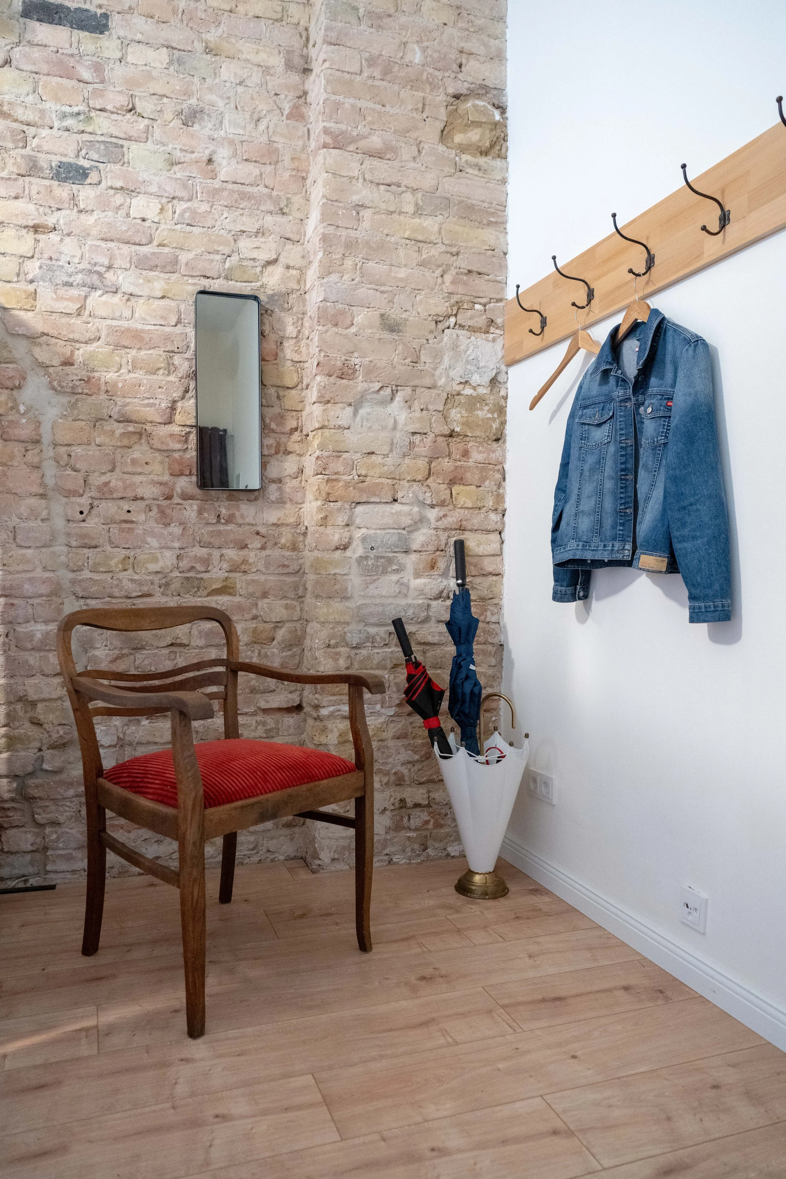 Ein Raum mit einer roten Polsterstuhl, einer Steinmauer, einem zusätzlichen weißen Wandabschnitt, einem Wandspiegel, einer Holzleiste mit vier Haken, an einem Haken hängt eine Jeansjacke, daneben stehen Regenschirme in einem weißen Behälter, Steckerl