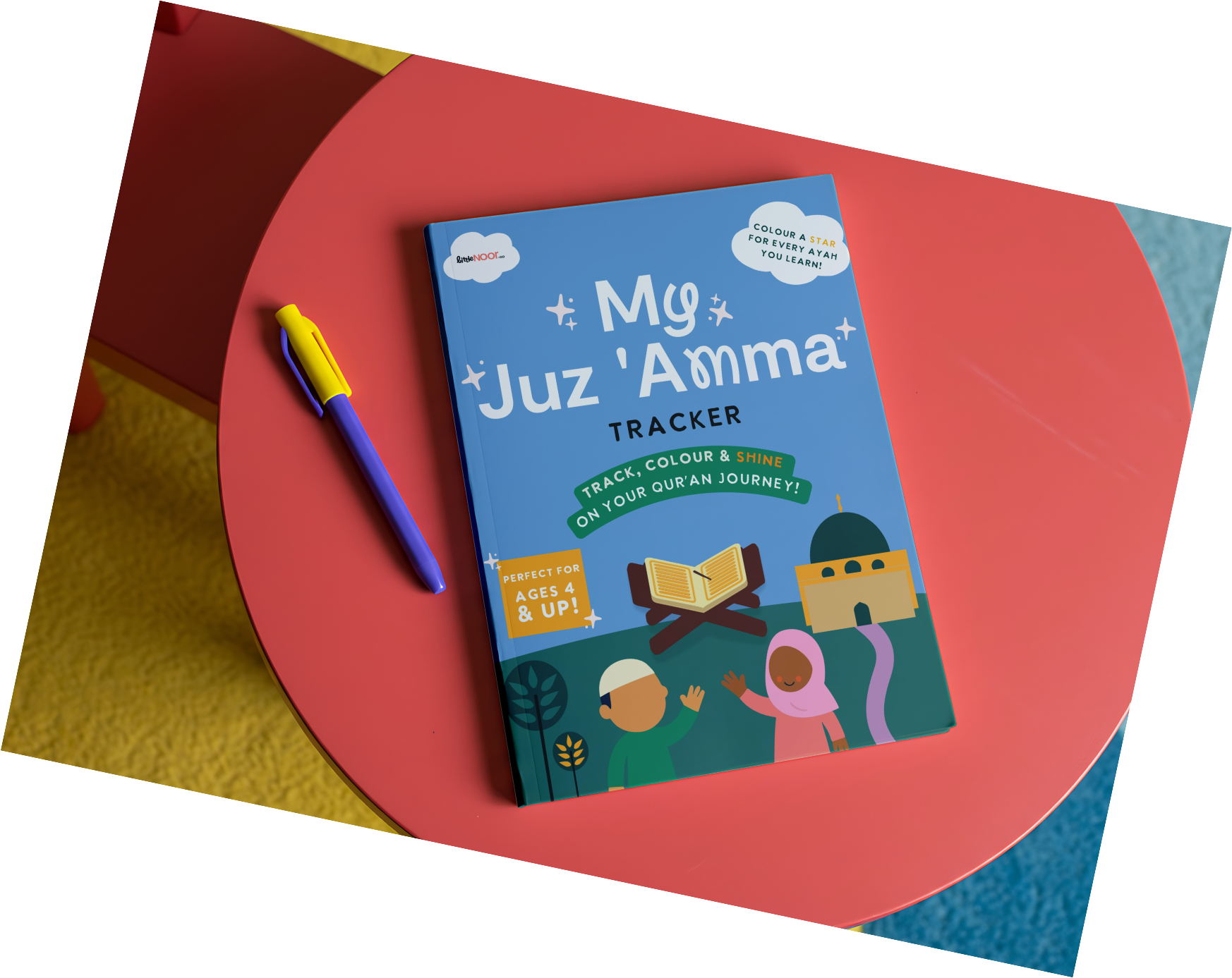A children's book titled 'Mg Juz 'Aama' Tracker is placed on a pink table with a yellowish textured surface underneath, next to a blue and yellow pen.