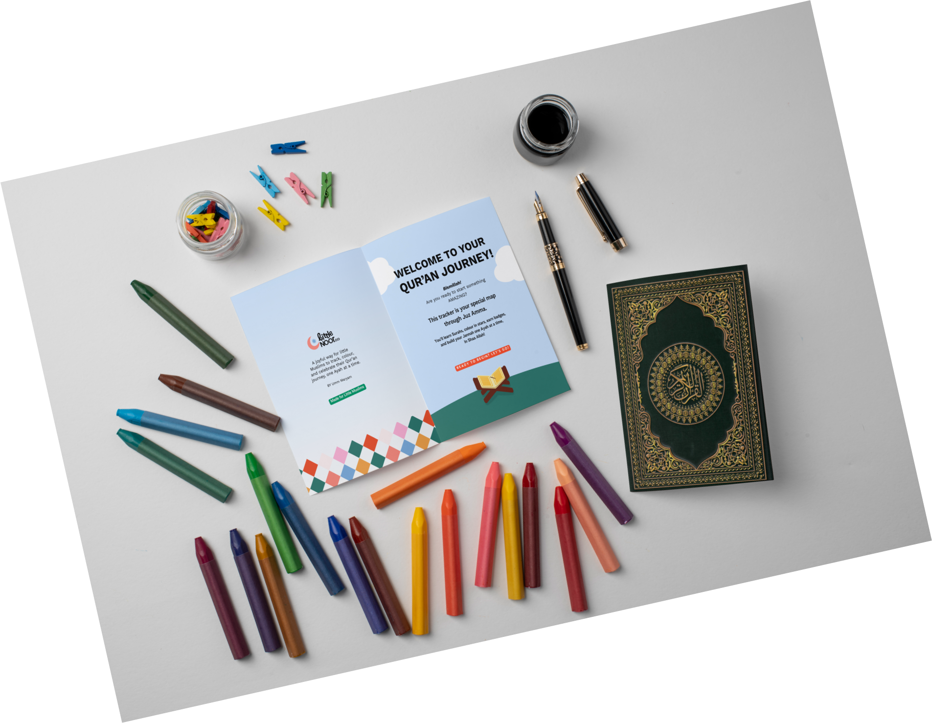 Colorful markers arranged in a semi-circle on a white surface with a Quran and a flyer, along with clothespins, a cup of ink, and a pen.