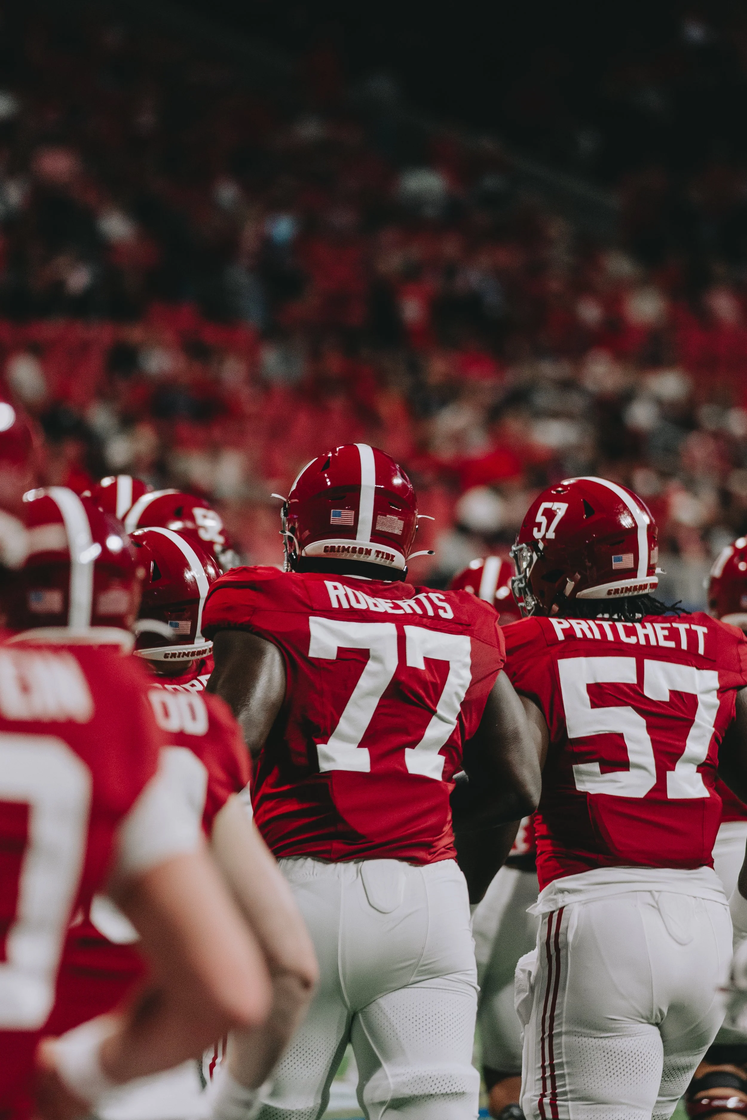 Alabama Crimson Tide football players in red jerseys and white pants standing in a huddle during a game.