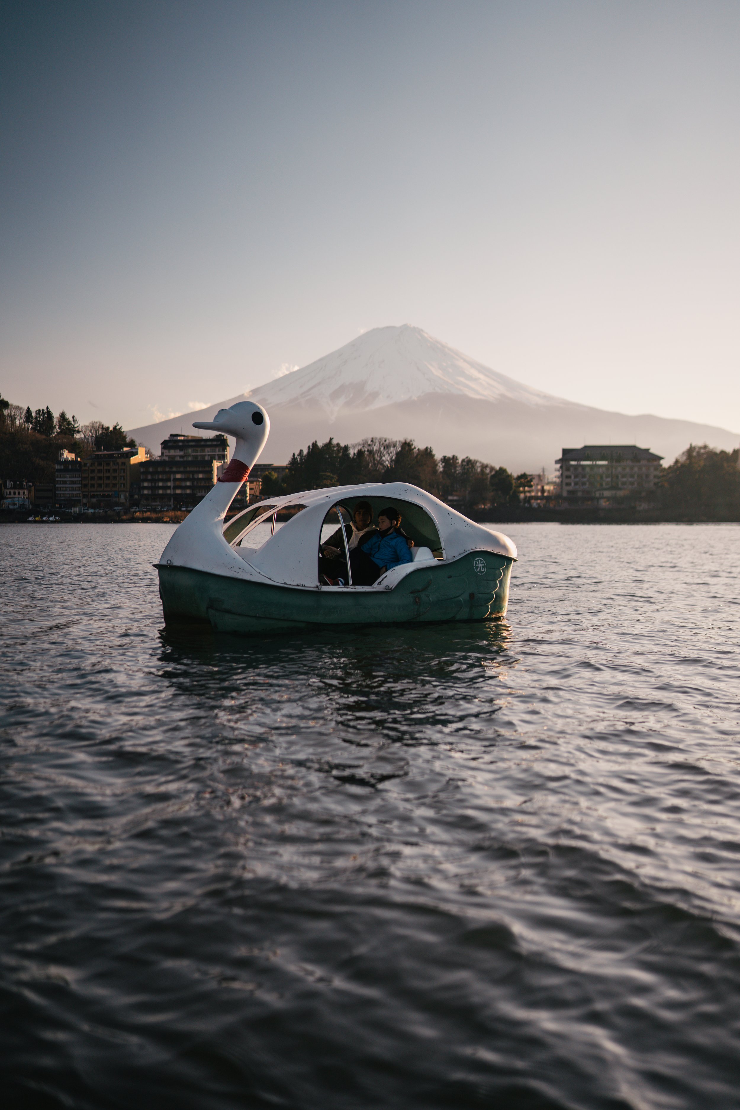 A swan-shaped pedal boat with two children on board floating on a lake, with Mount Fuji in the background during sunset.