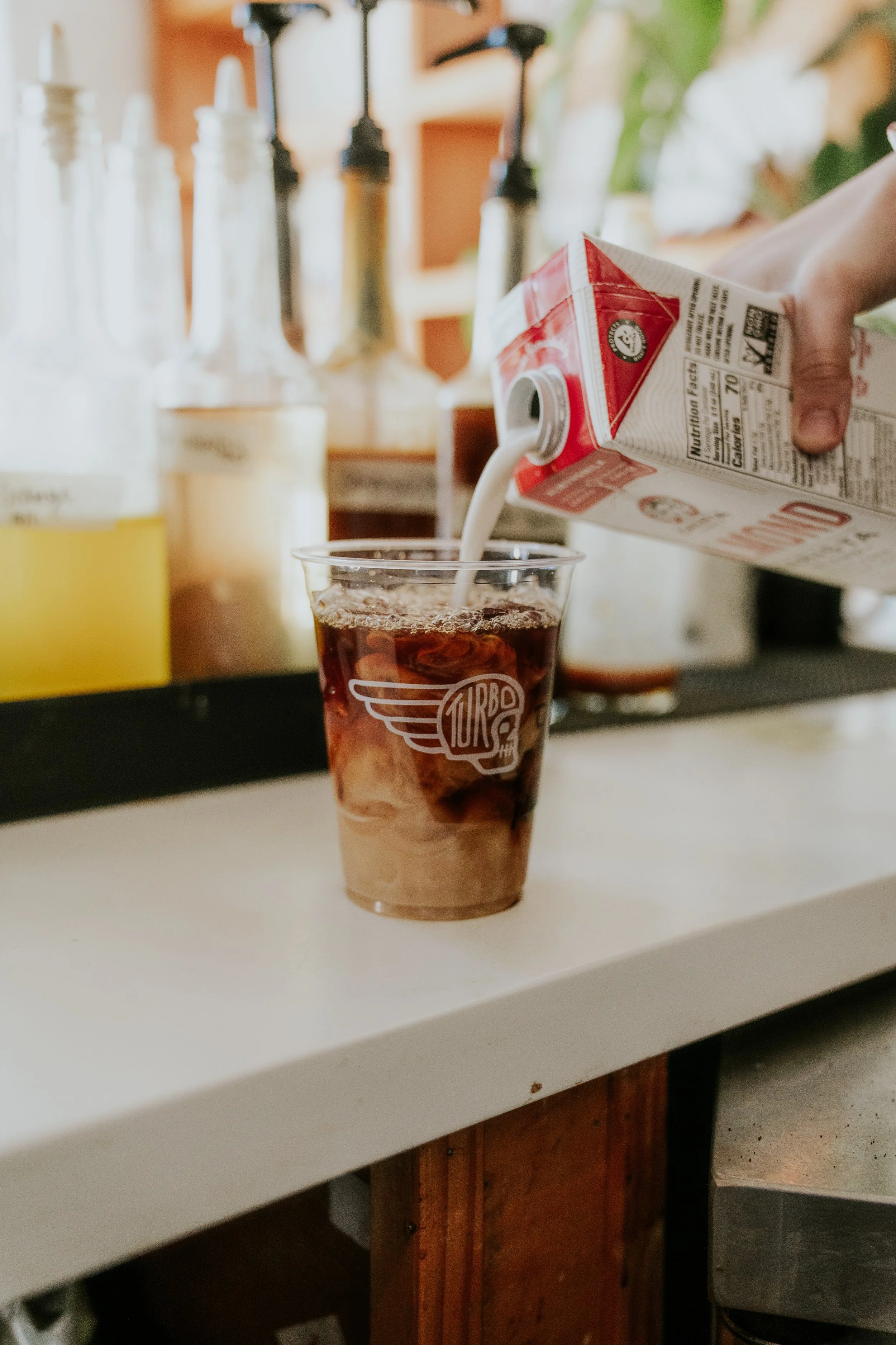A hand pouring milk into a glass of dark soda on a white countertop, with various bottles and kitchen items in the background.
