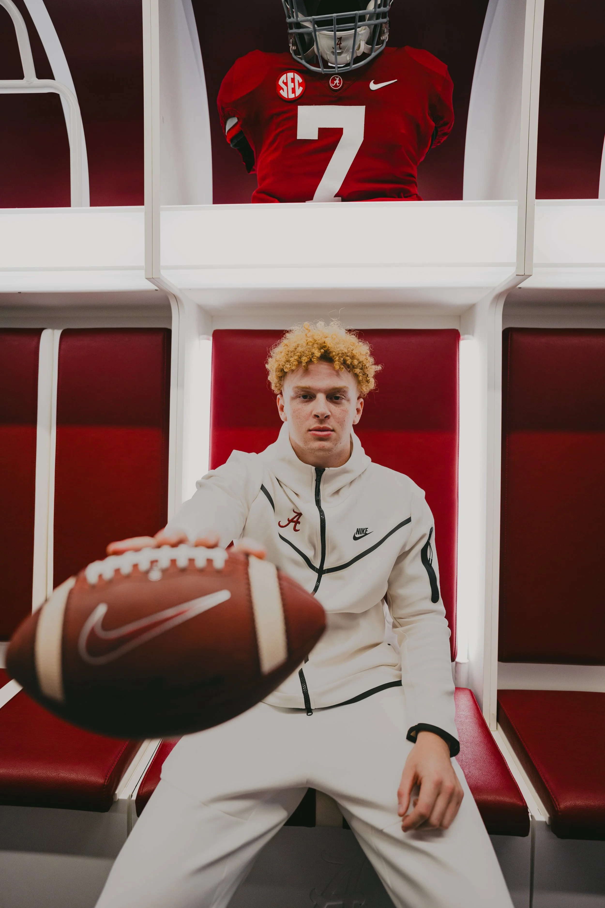 Young male athlete with curly red hair sitting on a red bench in a sports stadium, reaching out towards the camera with a football. He is dressed in a white sports jacket with black accents, and his face is focused and intense.