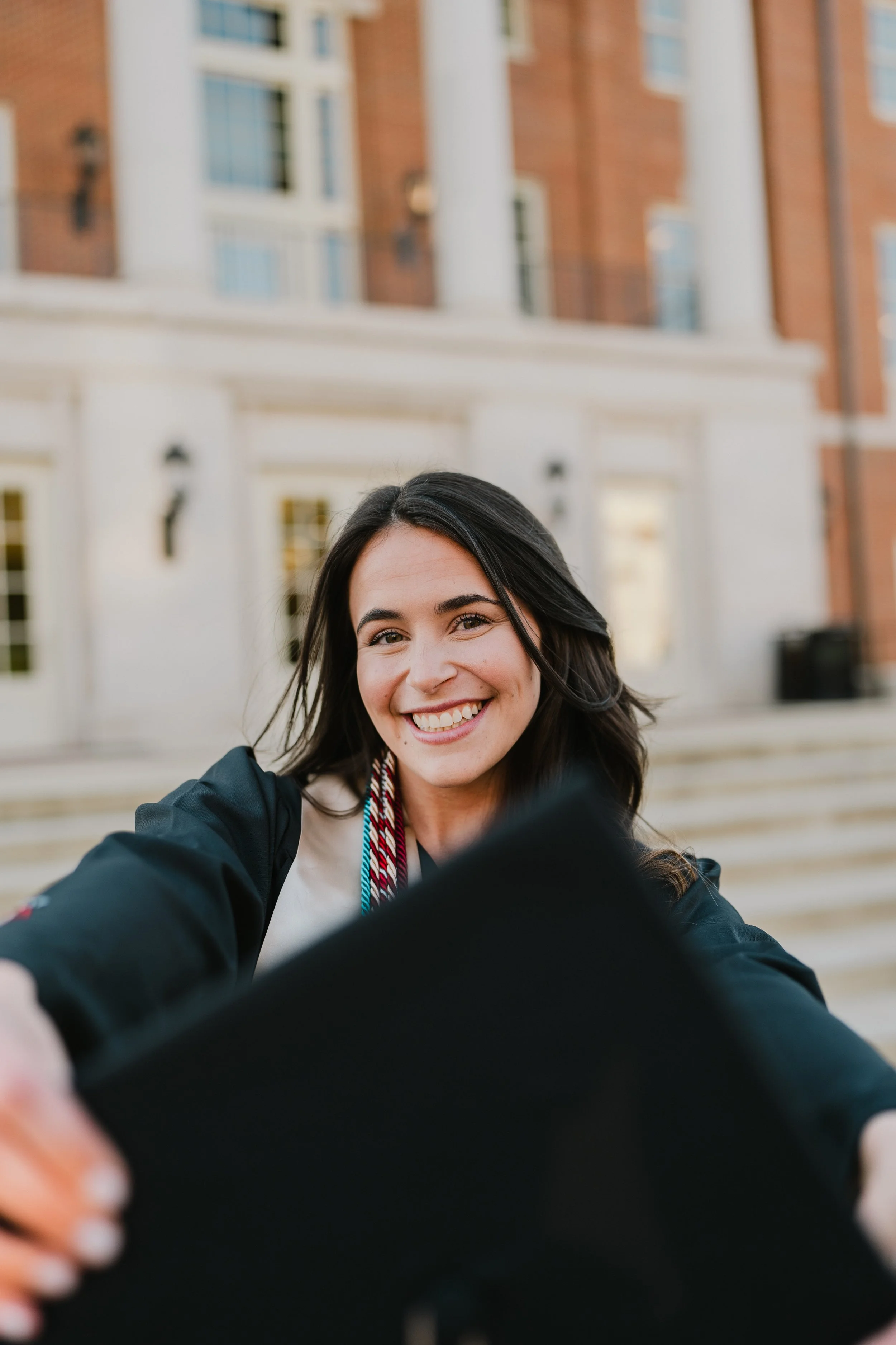 A young woman with long dark hair smiling and taking a selfie in front of a building with steps and columns.