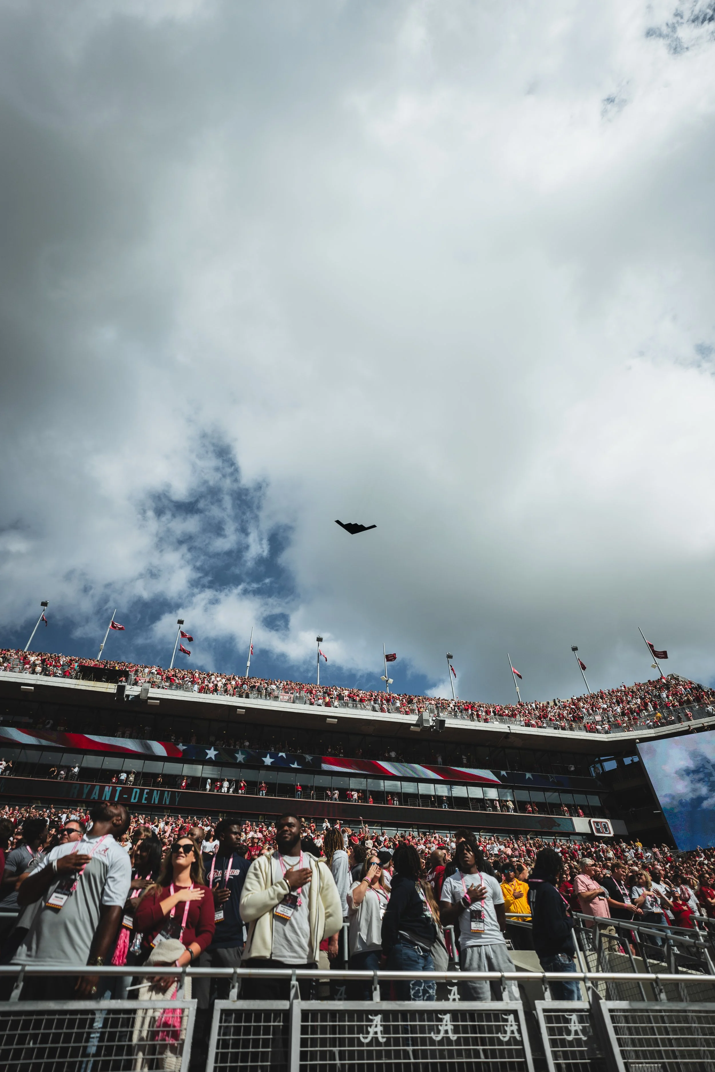 Crowd of people standing and watching a sports event at a stadium with American flags and a partly cloudy sky.