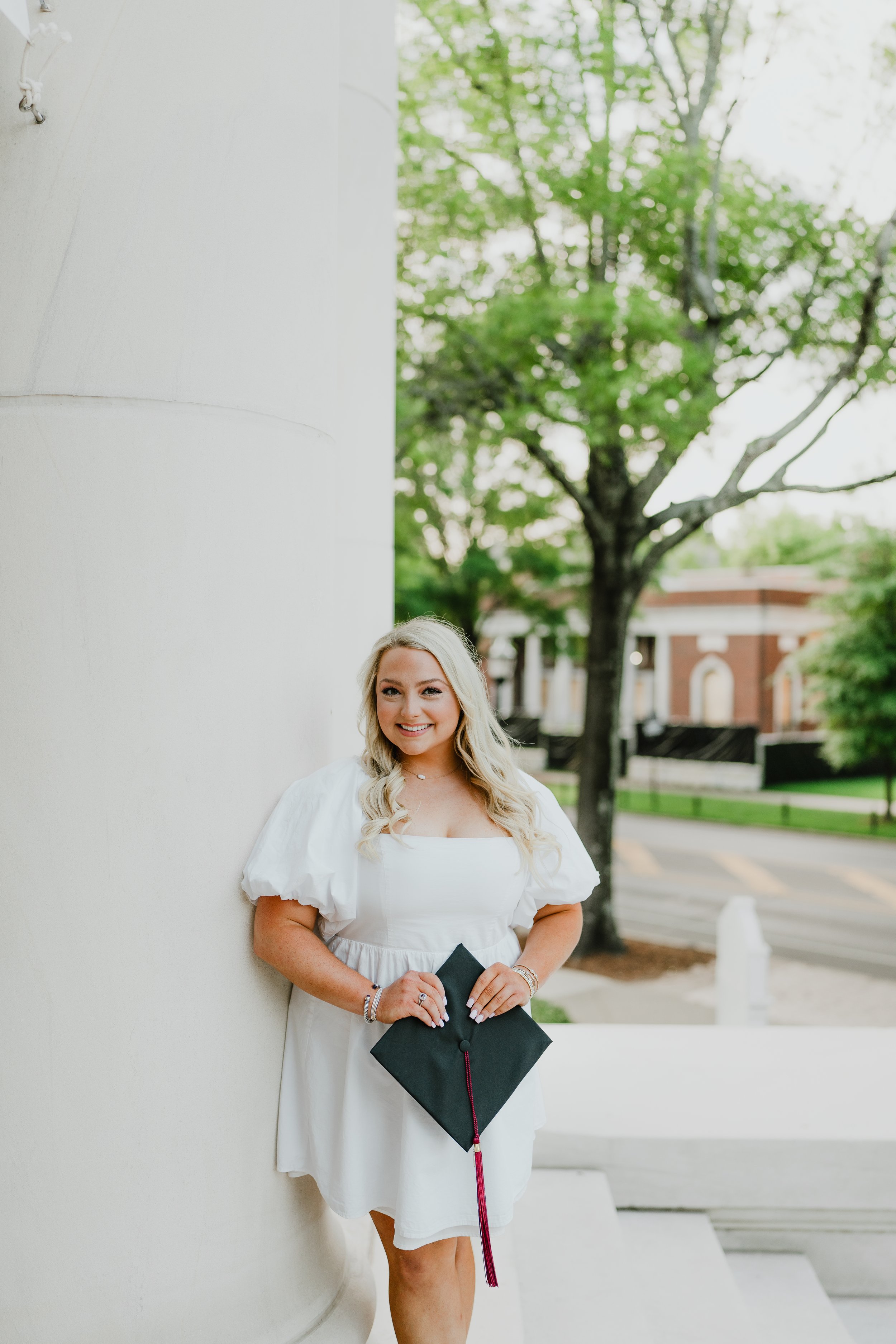 A woman with blonde hair, wearing a white dress, holding a black diploma cover, standing outdoors near a white wall with trees and a building in the background.