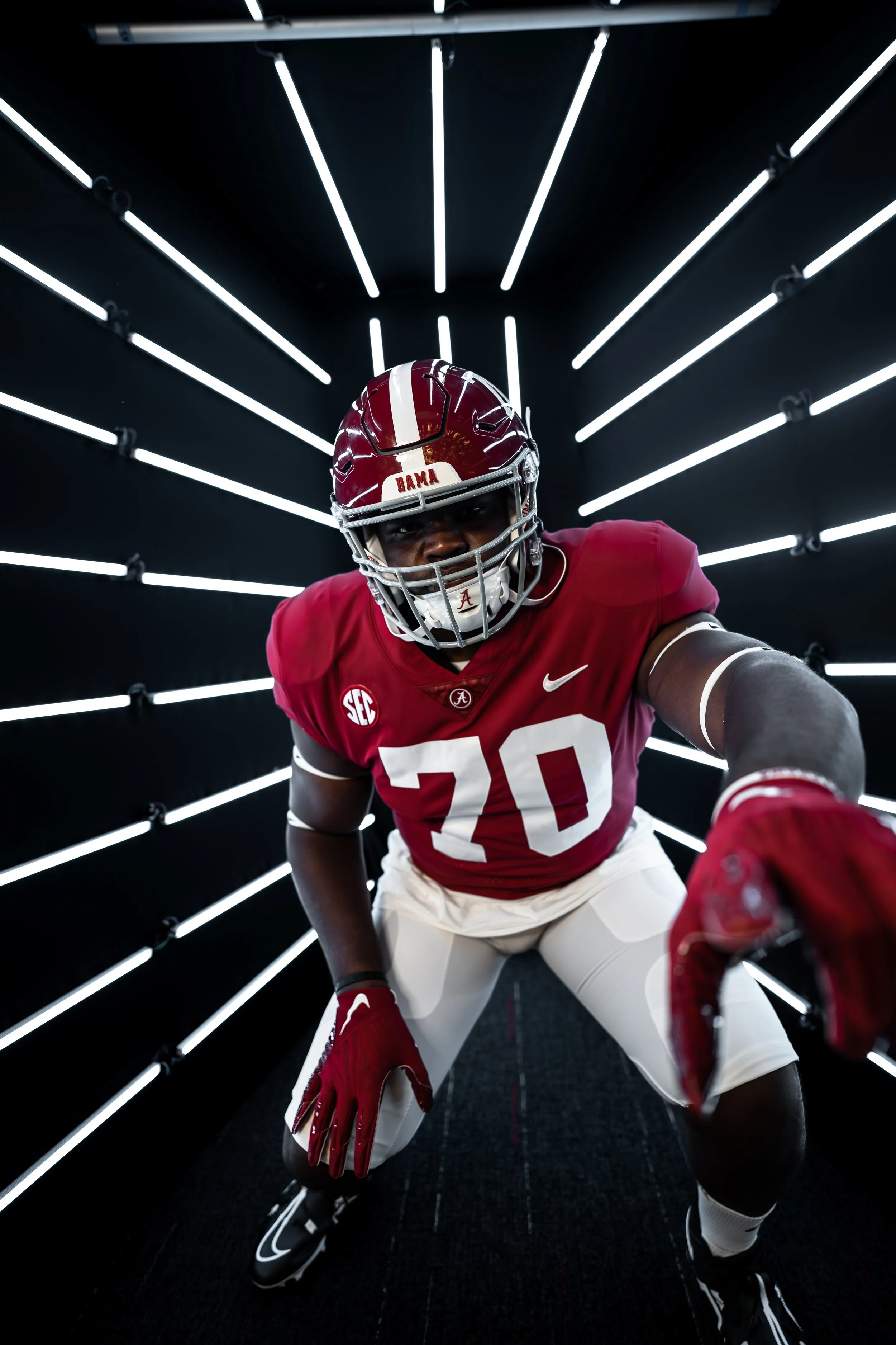 American football player in Alabama Crimson Tide uniform crouching in a futuristic black tunnel with white glowing light bars on the ceiling and walls.