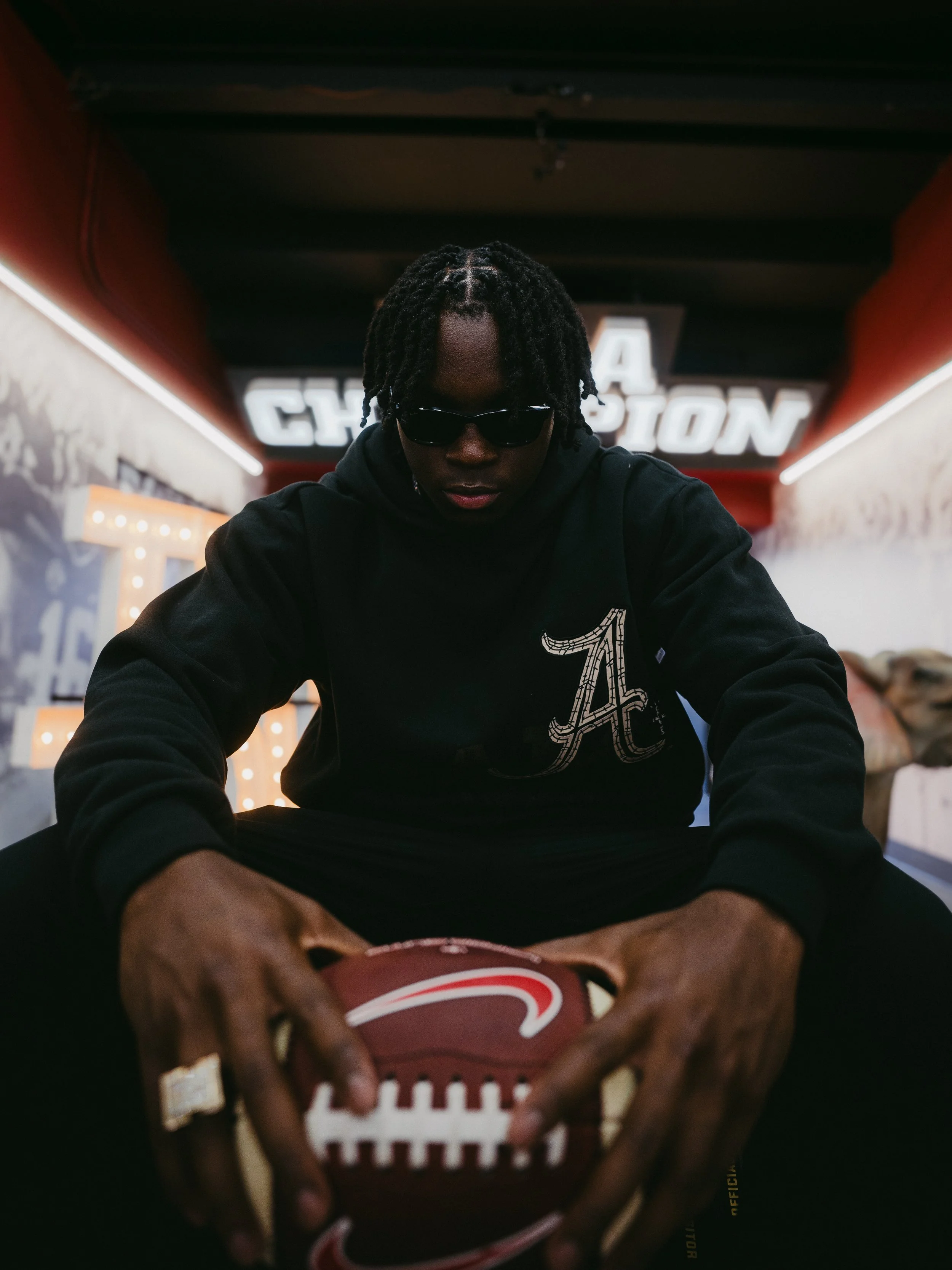A person with braids wearing sunglasses and a black hoodie, holding a football in a dimly lit setting with neon lights and signs in the background.