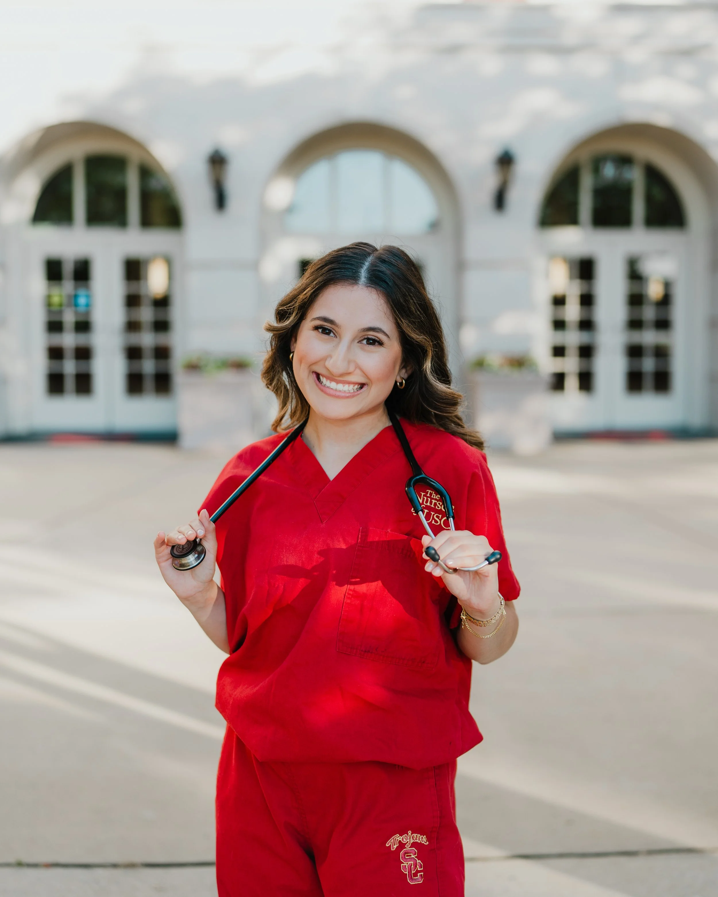 A young woman in a red medical scrub set holding a stethoscope, smiling, standing outside in front of a white building with large arched windows and black lanterns.