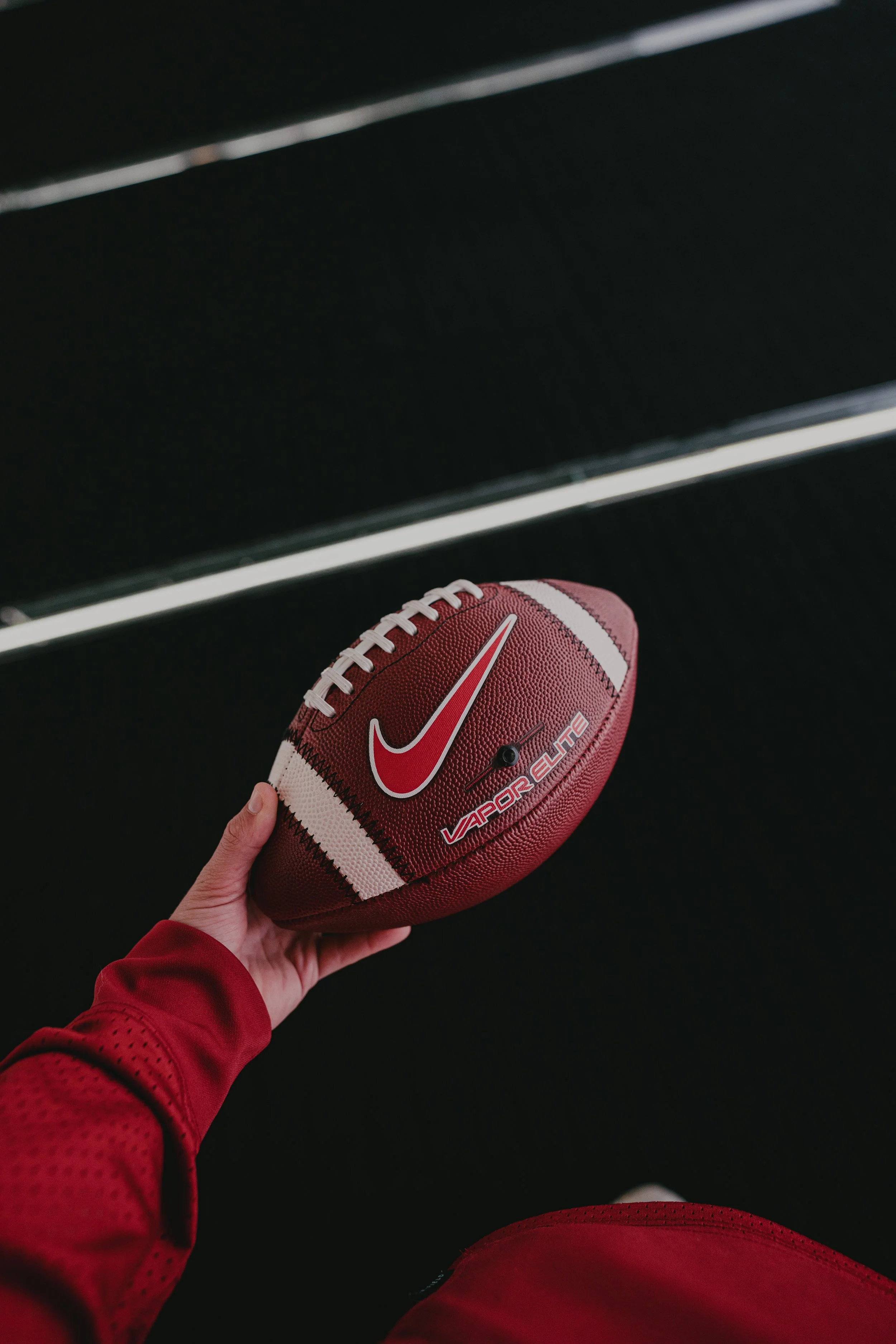 A person in red sportswear holding a red and white Nike football labeled "VAPOR ELITE" against a dark background.
