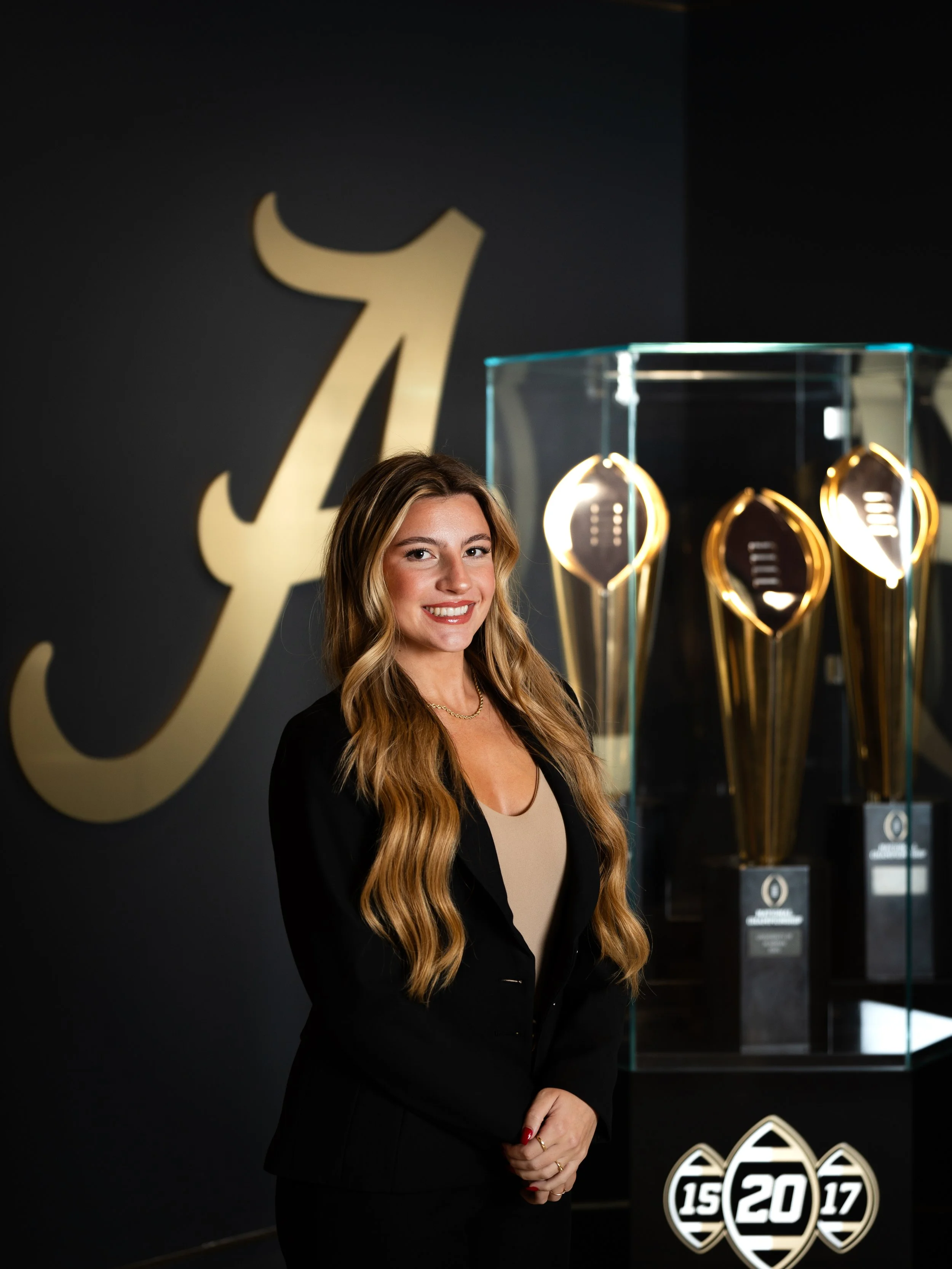 A woman with long, wavy blonde hair, wearing a black blazer and beige top, smiling and standing in front of a dark wall with a large logo and trophy display celebrating a sports achievement from 2015, 2017, and 2020.