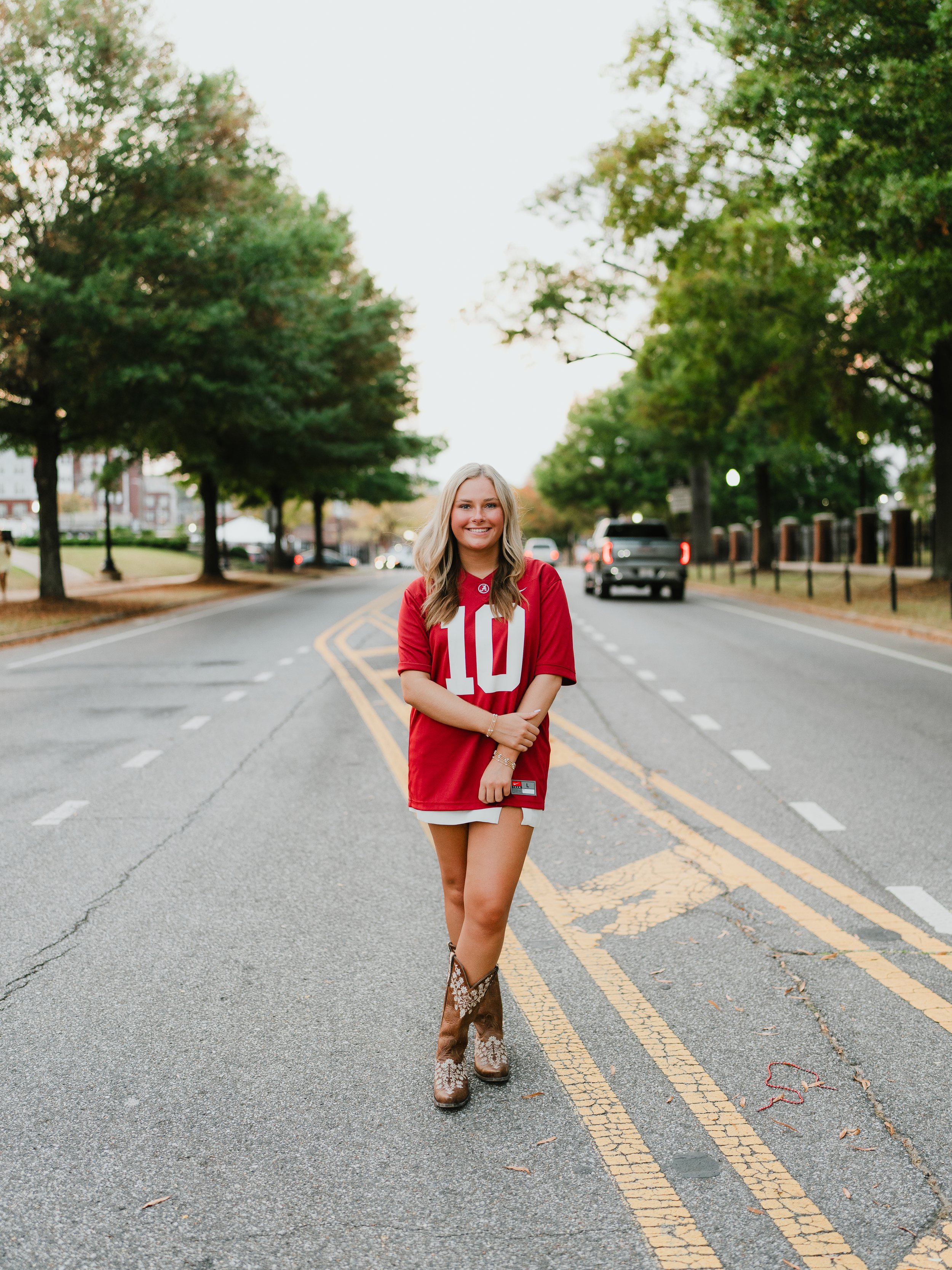A young woman with blonde hair smiling and wearing a red sports jersey and cowboy boots, standing on a road surrounded by trees, with vehicles in the background.